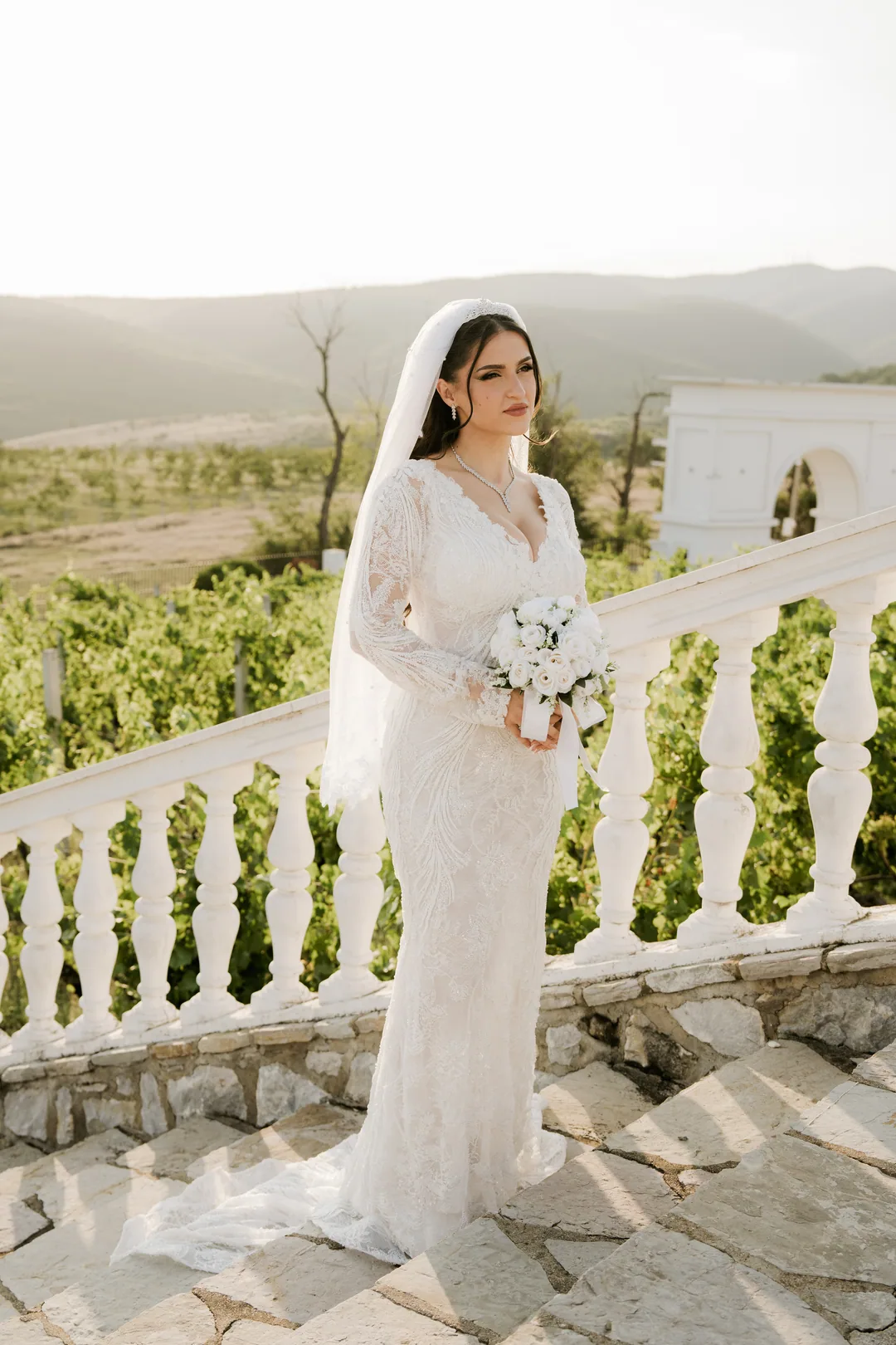 Bride alone on the balustrade steps with her bouquet, vineyard and hills beyond