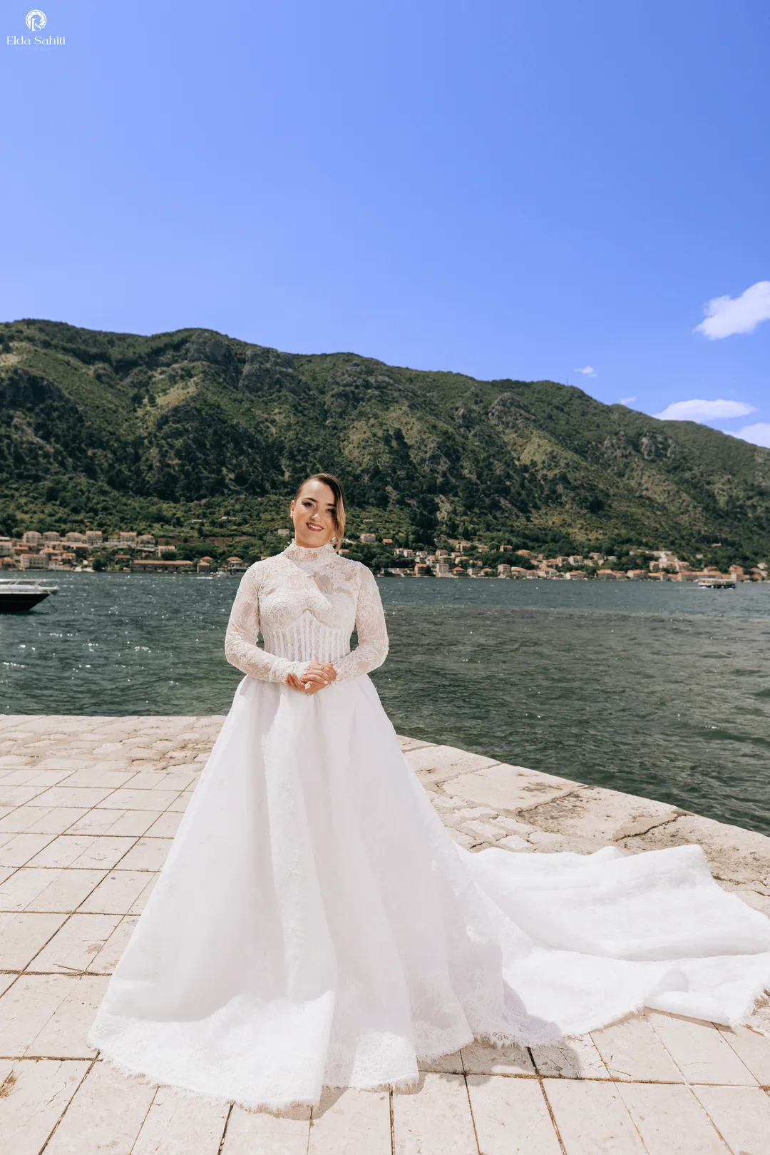 Bride standing alone on the quay, her train spread across the stone, mountains beyond