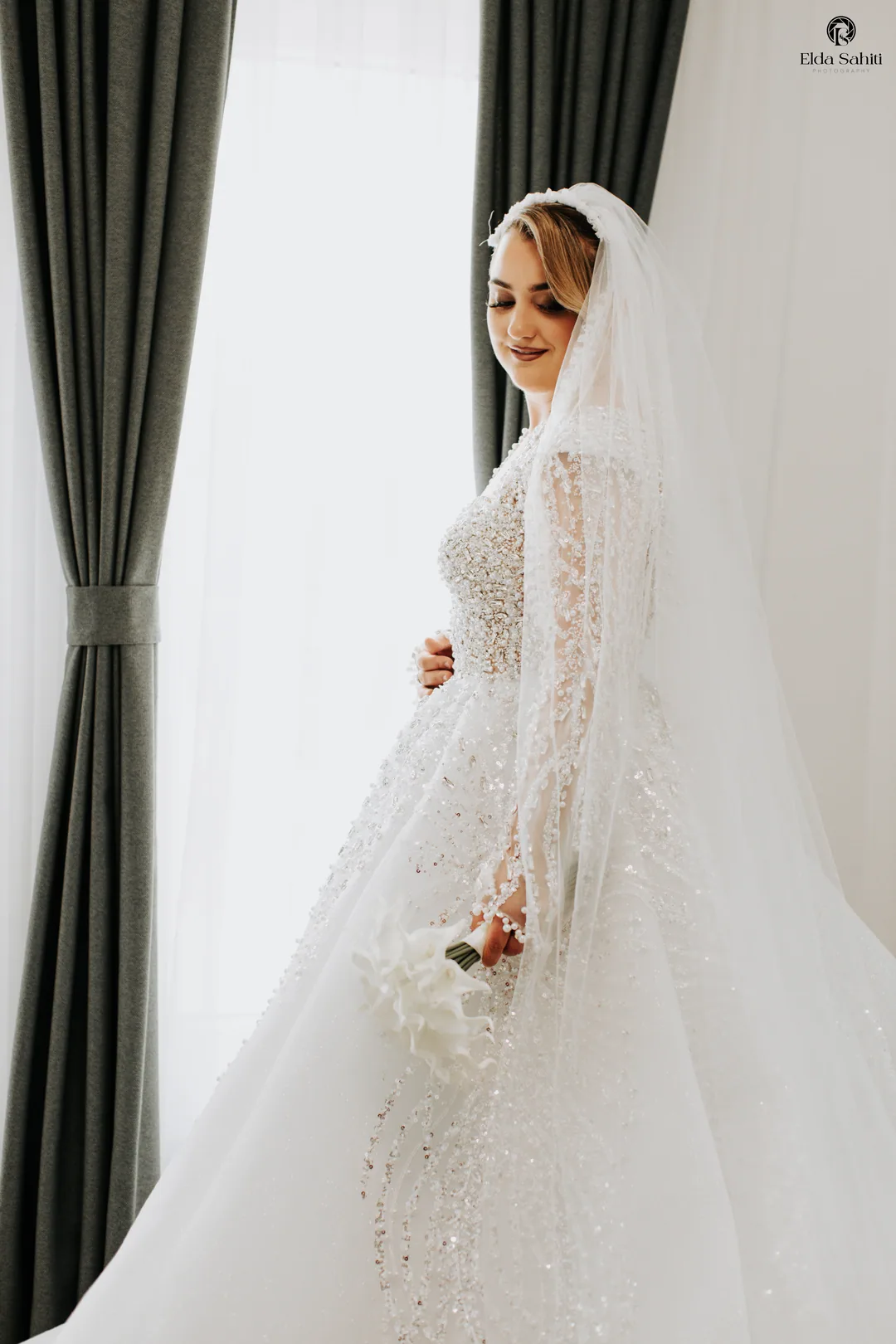 Bride in a beaded gown smiling down by a window with grey curtains