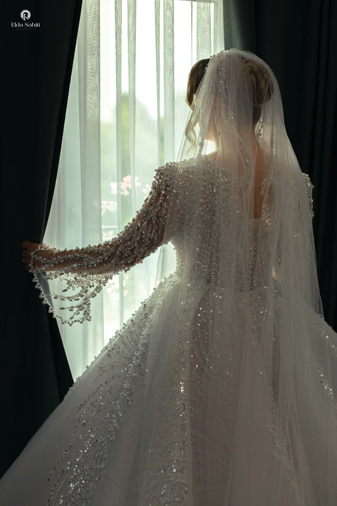 Bride from behind at the window, one hand on the curtain, her veil falling down her back