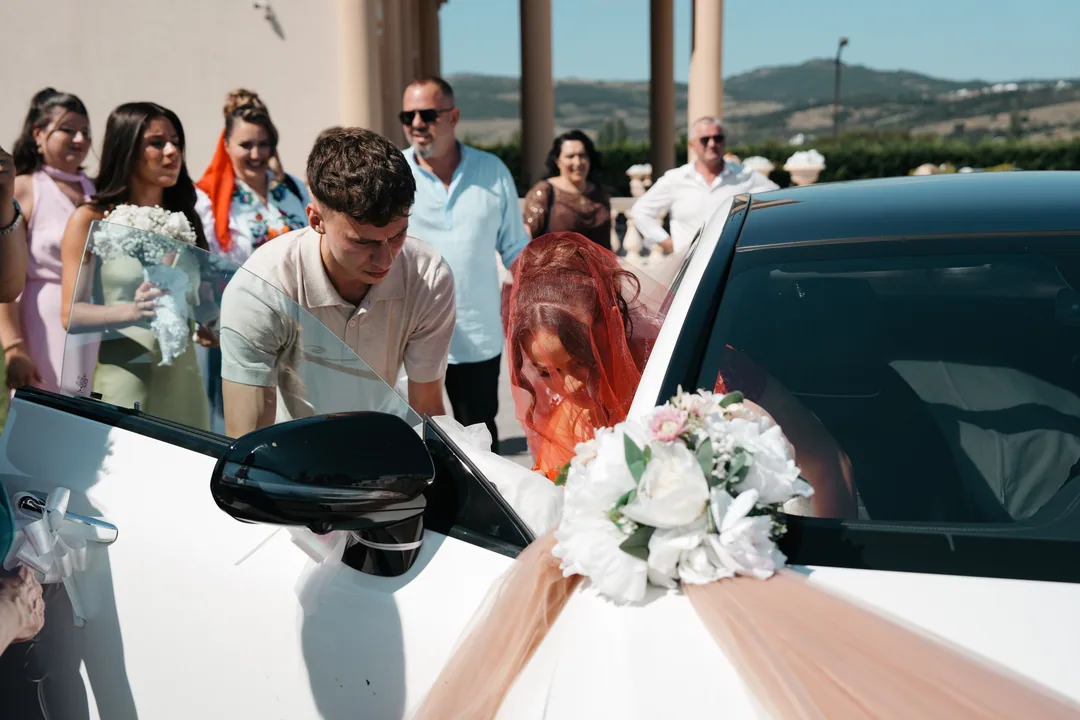 Bride in her red veil getting into the flower-decked wedding car as guests gather