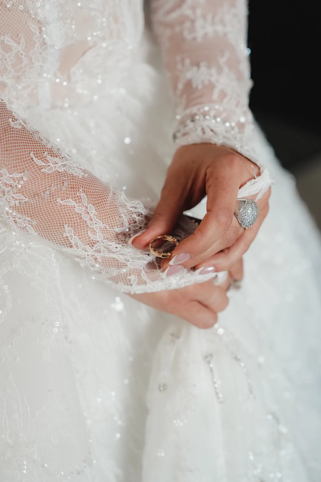 Bride holding a gold ring between her fingertips, lace sleeves over the embroidered skirt