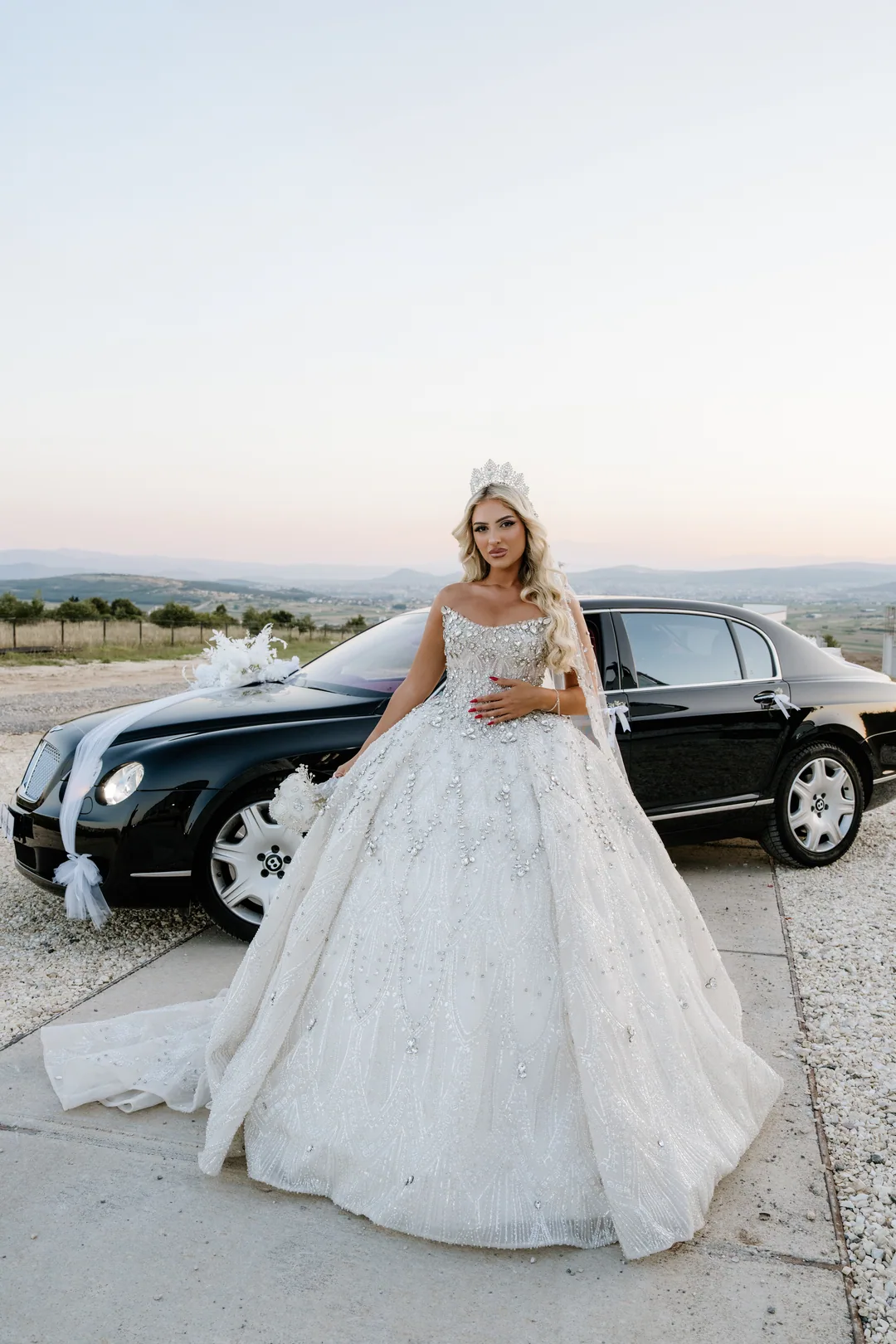 Bride in a crystal ball gown in front of the black car, hills behind