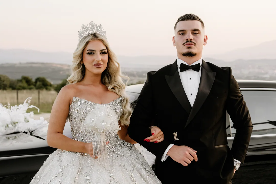Bride in a crown and groom arm in arm, looking into the camera, hills on the horizon