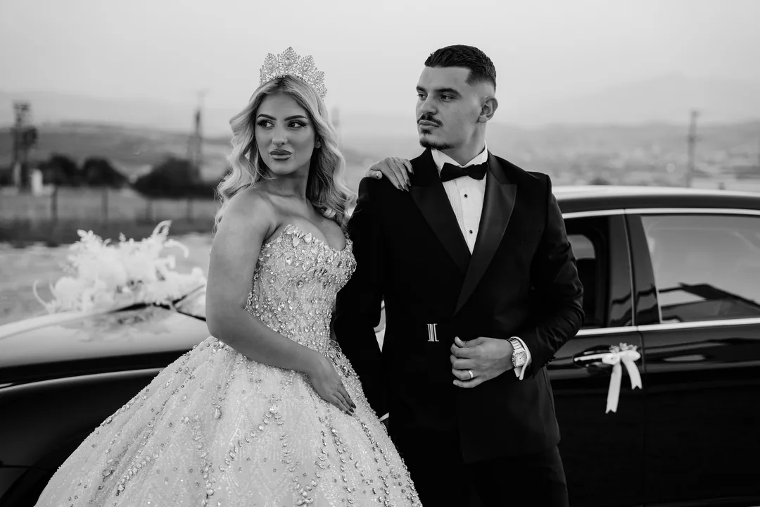 Bride in a crown glancing at the groom beside the black car, in black and white