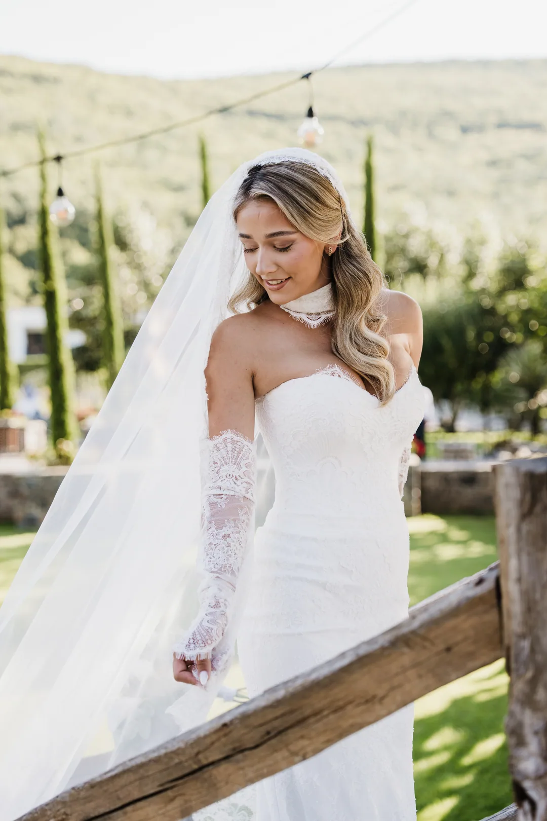 Bride in lace gloves leaning on a wooden fence, smiling down, string lights above