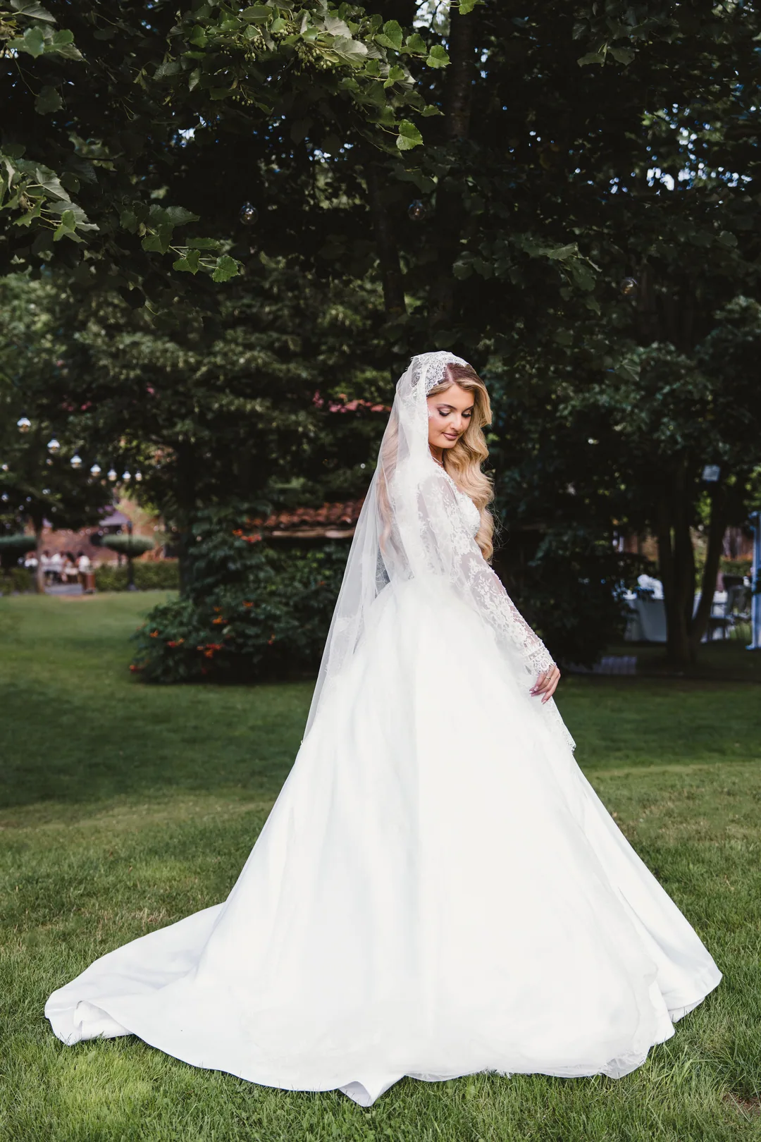 Bride in a long lace veil looking down, her skirt spread over the lawn