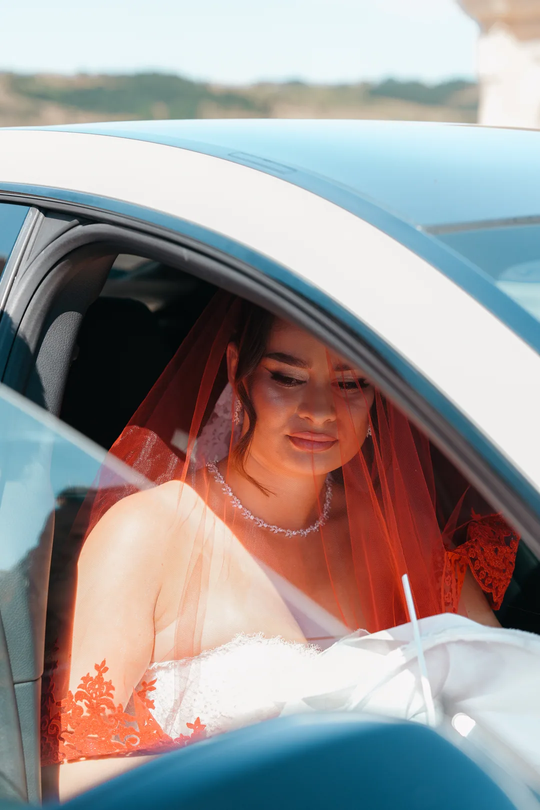 Bride under her red veil sitting in the car, looking down with a quiet smile