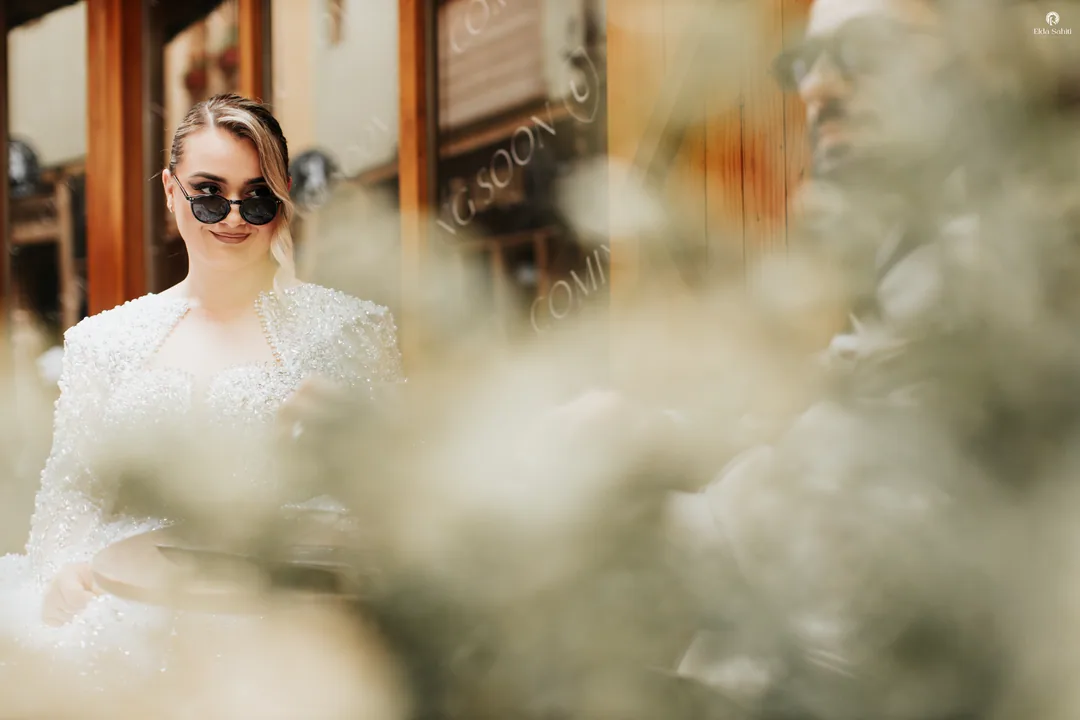 Bride in sunglasses at a café table, seen through blurred leaves