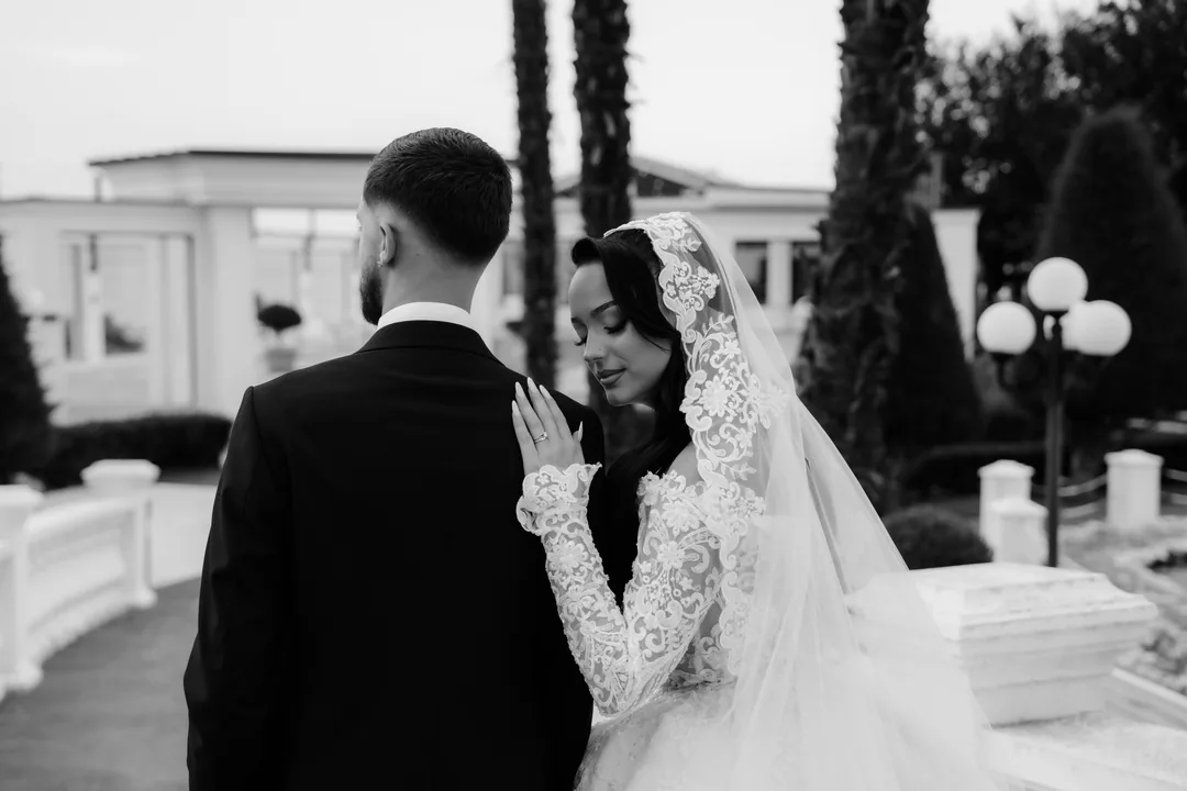 Bride resting her hand on the groom's shoulder outside a white palace, in black and white