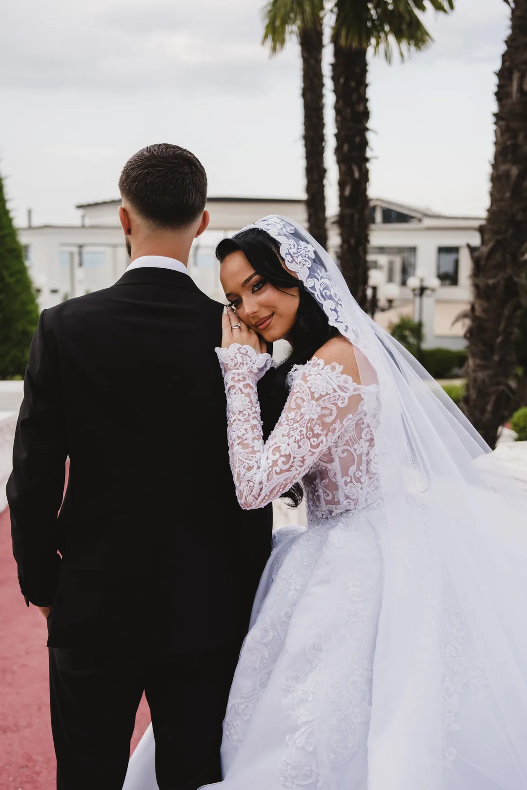 Bride in a lace mantilla looking over the groom's shoulder, palm trees behind