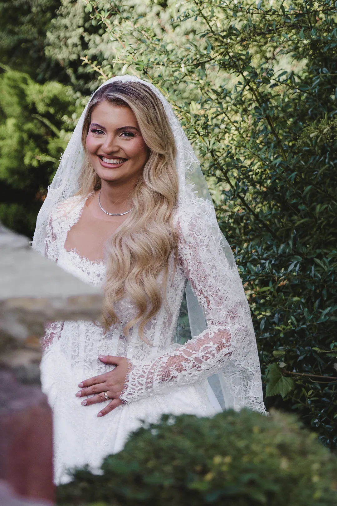 Bride smiling beside a hedge, long waves under her lace veil