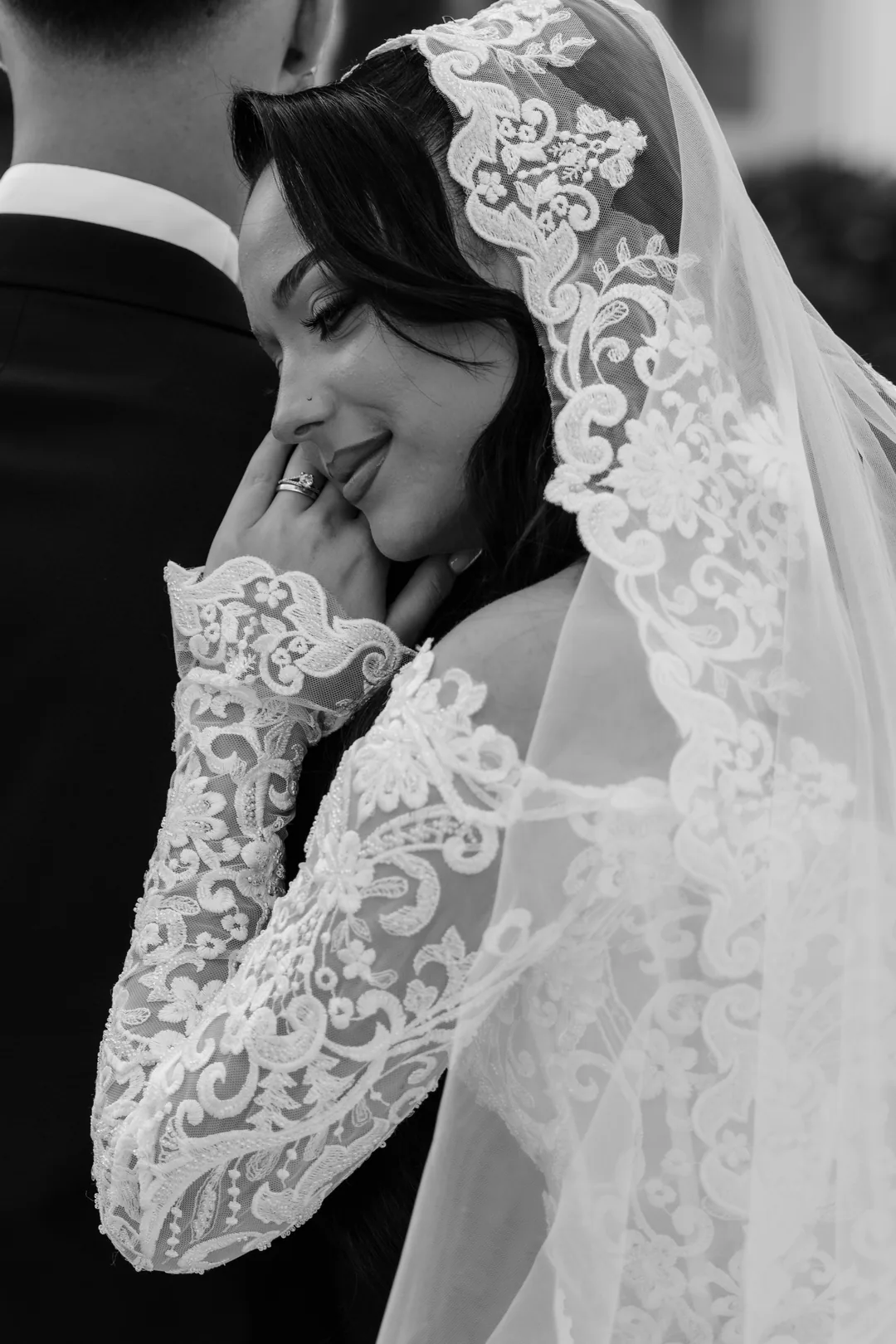 Bride smiling with her hand to her cheek, the lace veil framing her face, in black and white