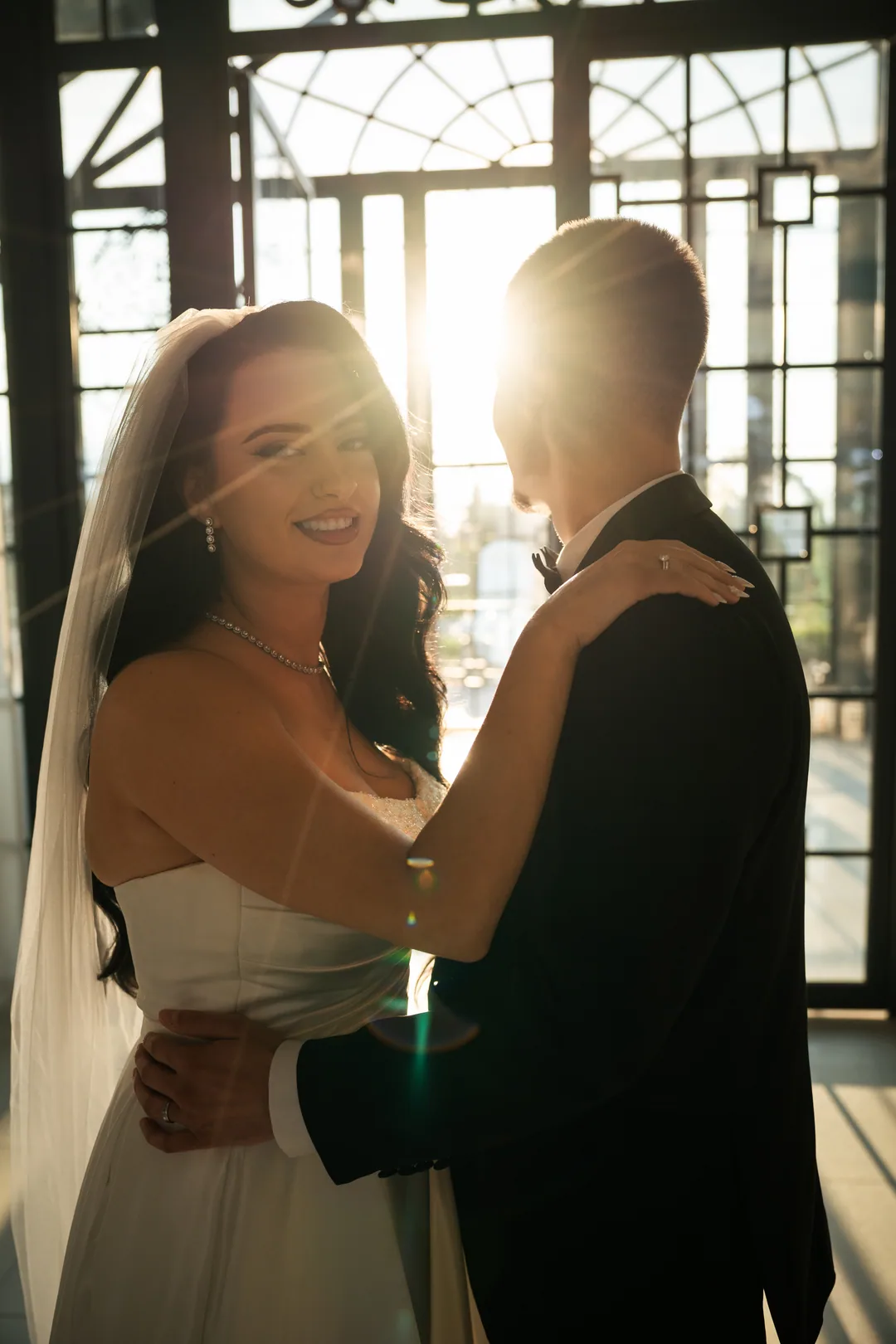 Bride smiling over the groom's shoulder as they embrace against the light