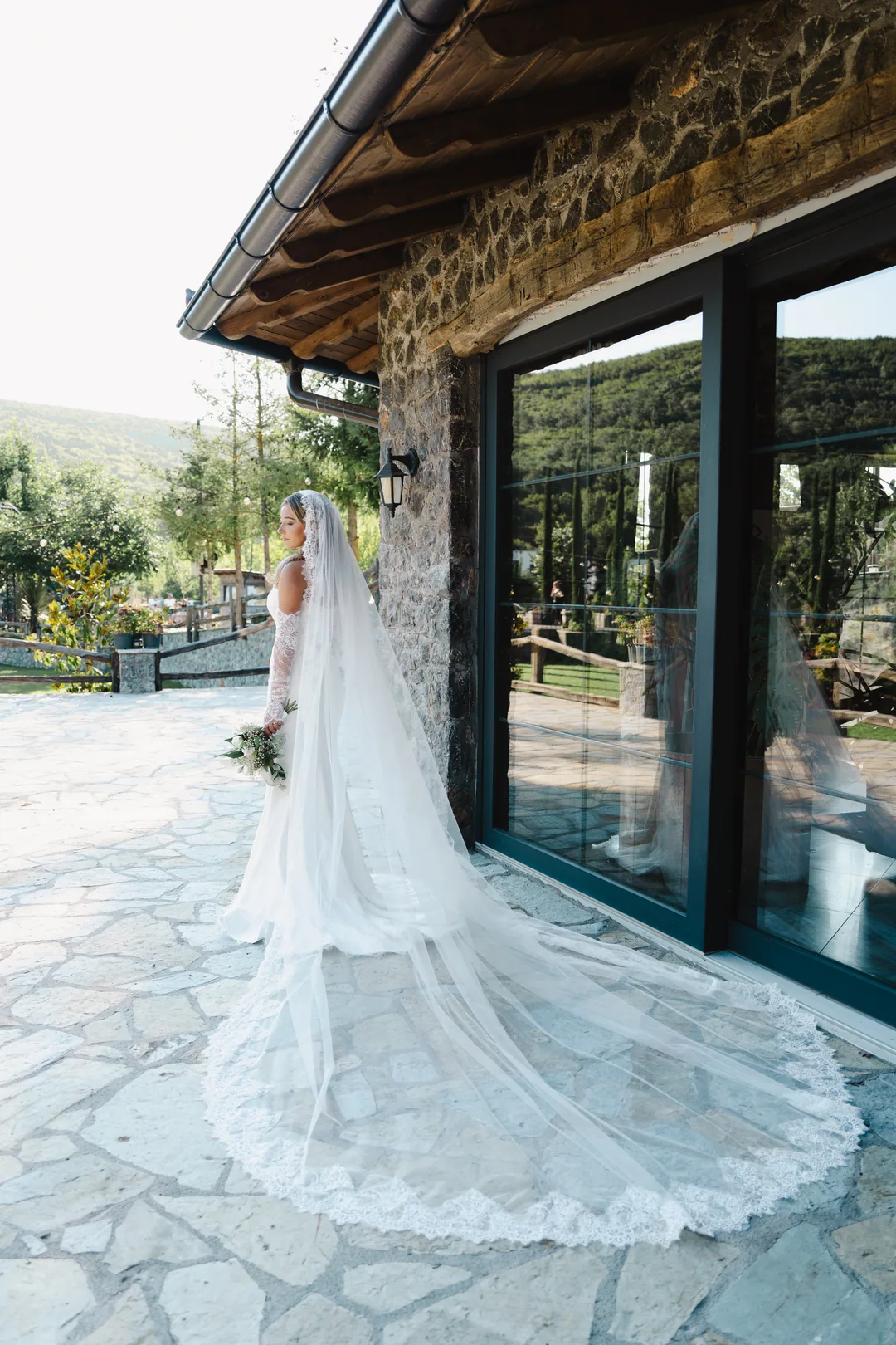 Bride from behind, her cathedral veil spread across the flagstones beside a stone house
