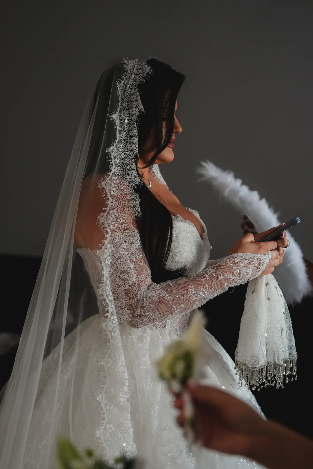 Bride in a lace veil checking her phone, a feather fan in her other hand