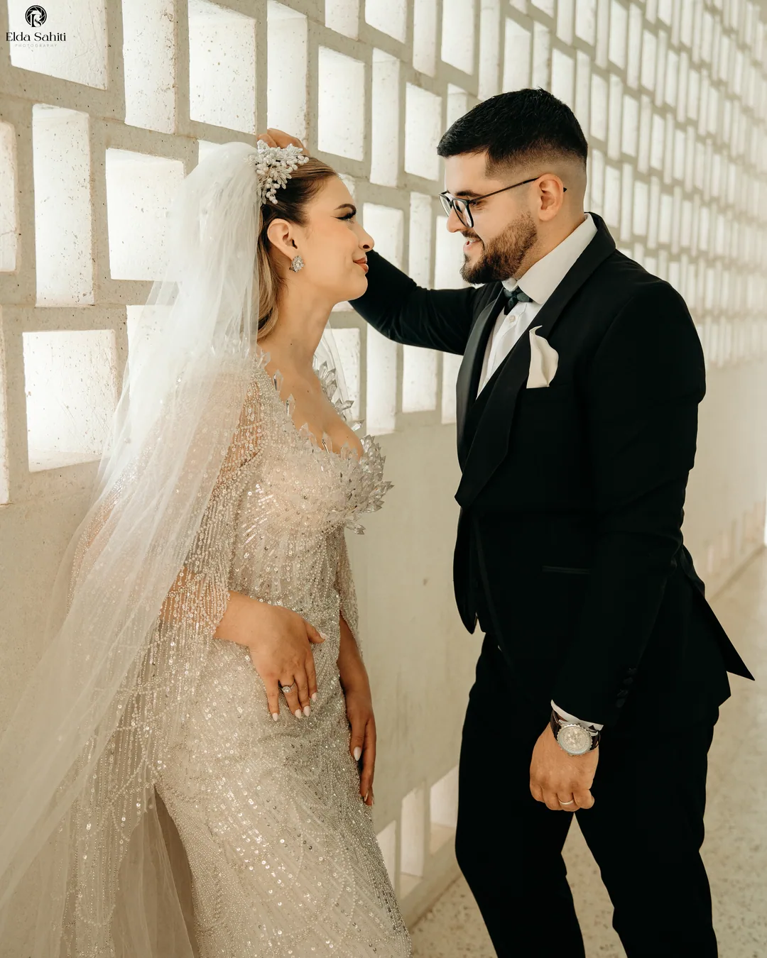 Groom touching the bride's hair against a white patterned wall, both smiling