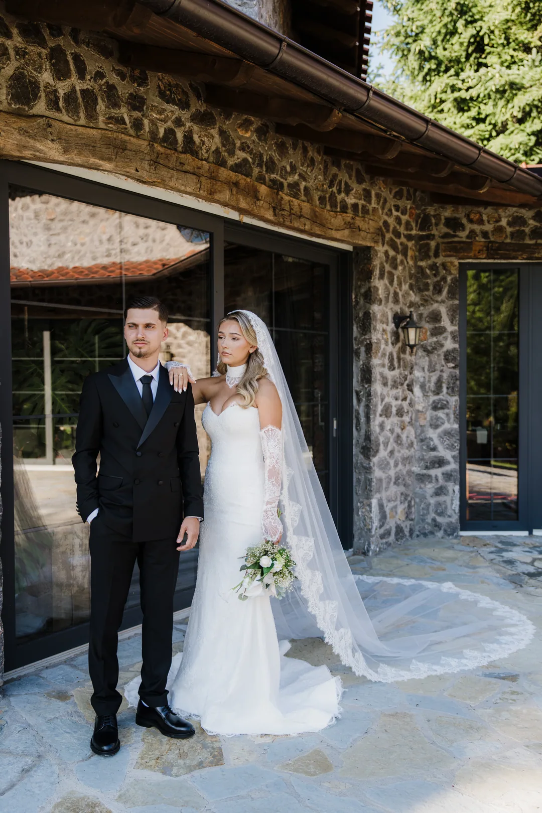 Bride and groom in front of the stone house, her veil fanned out behind her