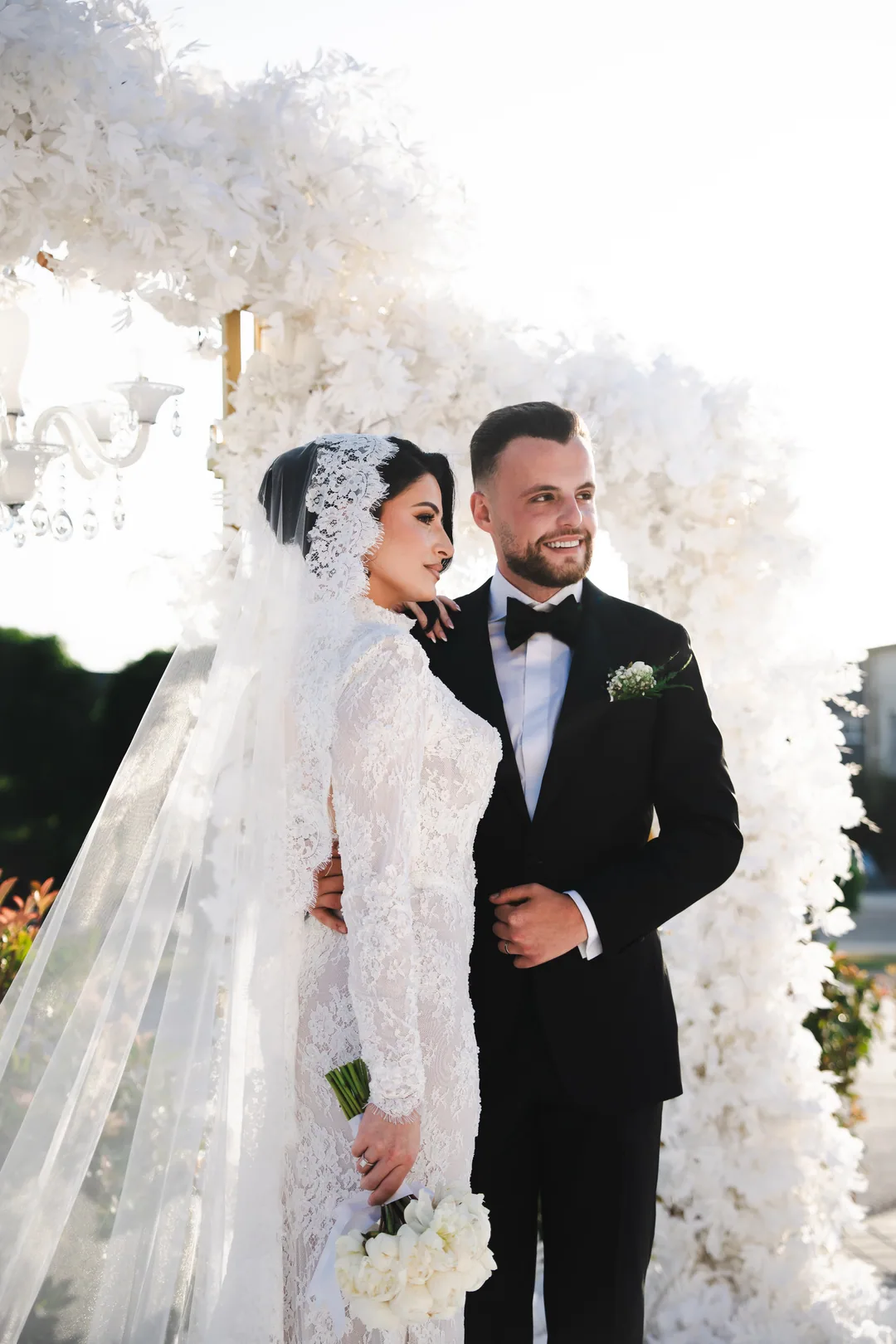 Bride in a lace headpiece leaning into the groom in front of a wall of white flowers