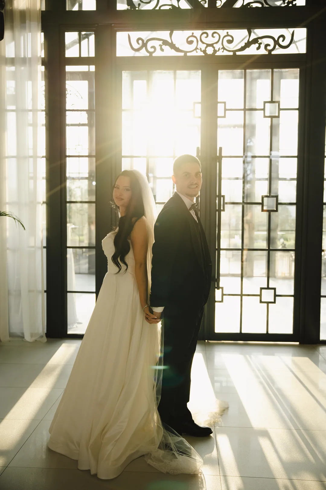 Couple back to back and smiling, sunlight pouring through the doors behind them