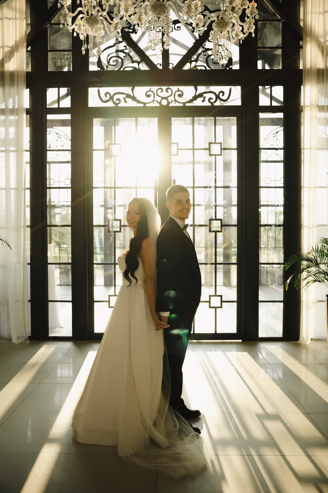 Bride and groom standing back to back in front of sunlit doors, long shadows on the floor