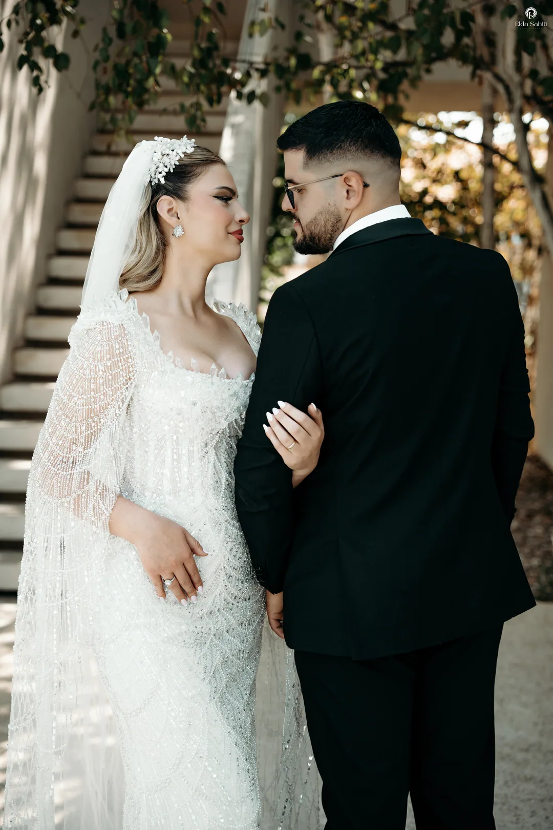 Bride and groom face to face at the foot of a white staircase, her lace sleeves catching the light