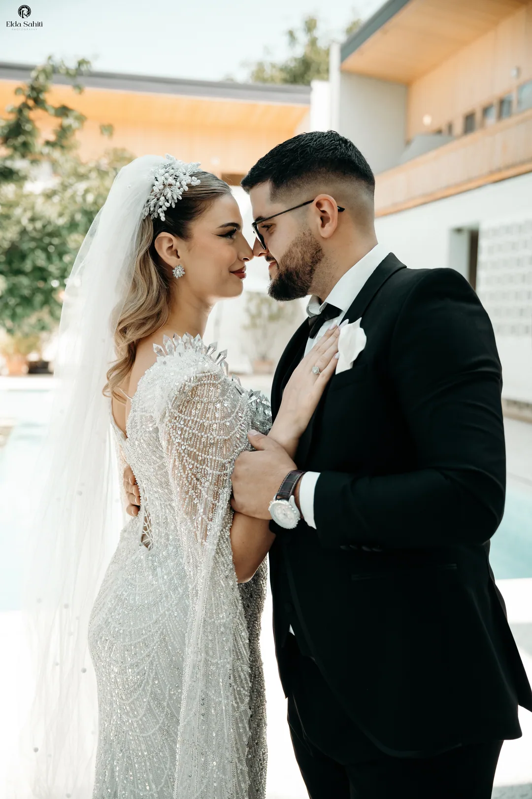 Couple forehead to forehead by the pool, her beaded cape sleeves falling to the ground