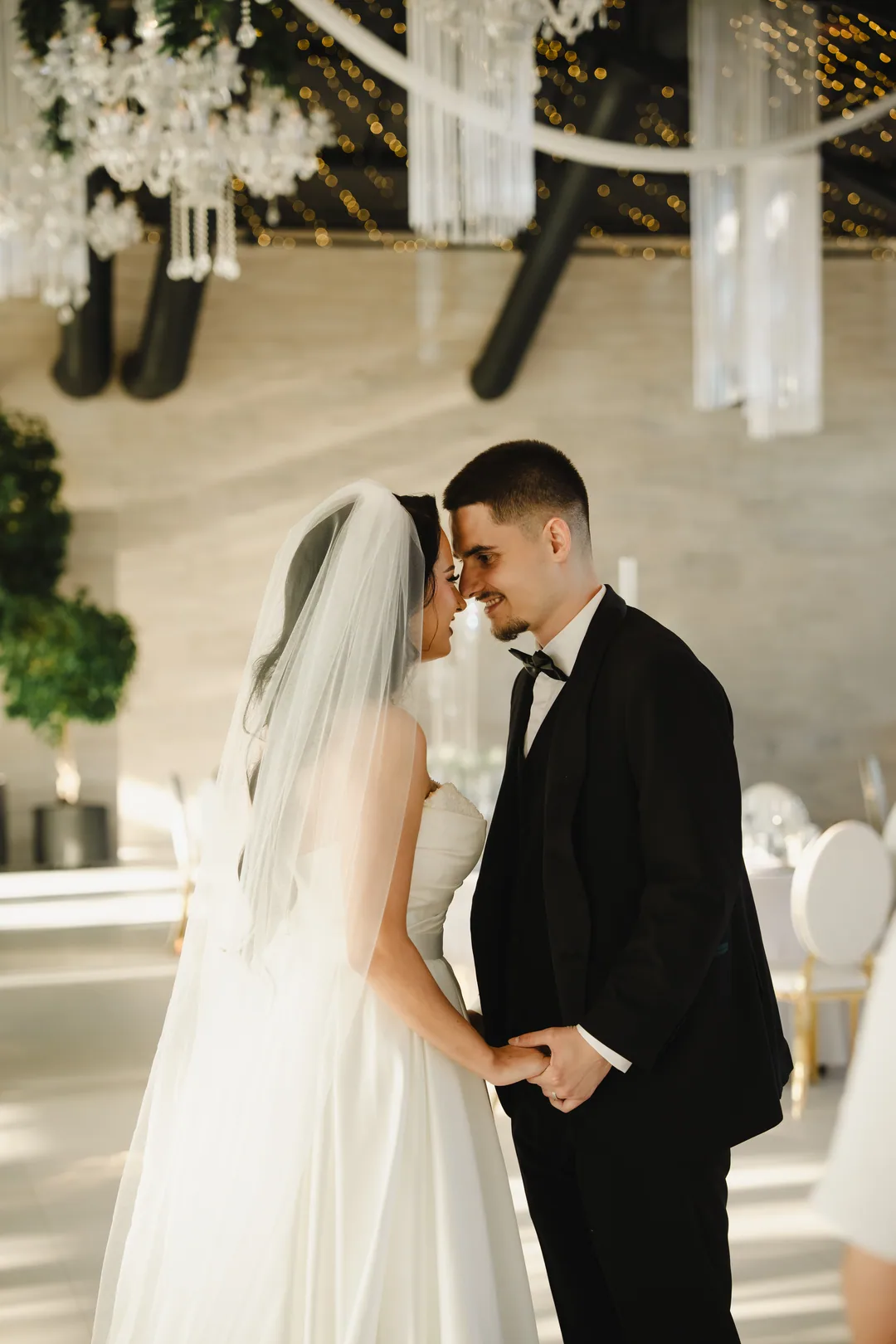 Couple forehead to forehead, holding hands, beneath crystal chandeliers