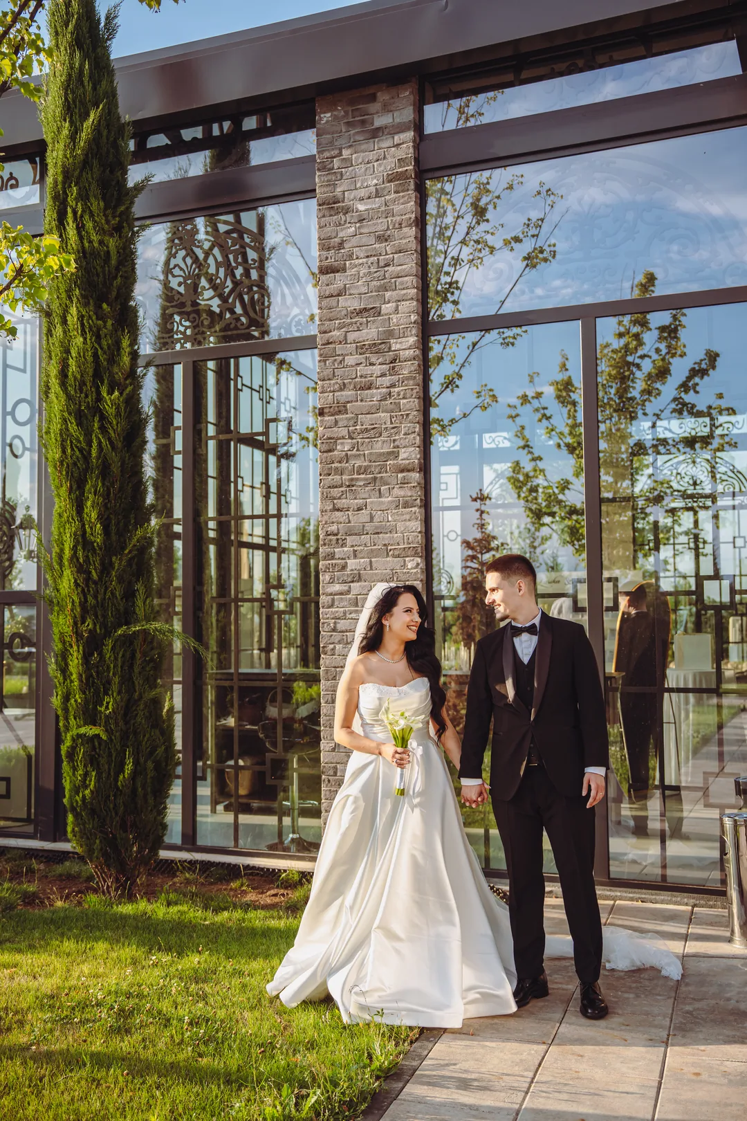 Bride and groom hand in hand outside the glass pavilion, a cypress beside them