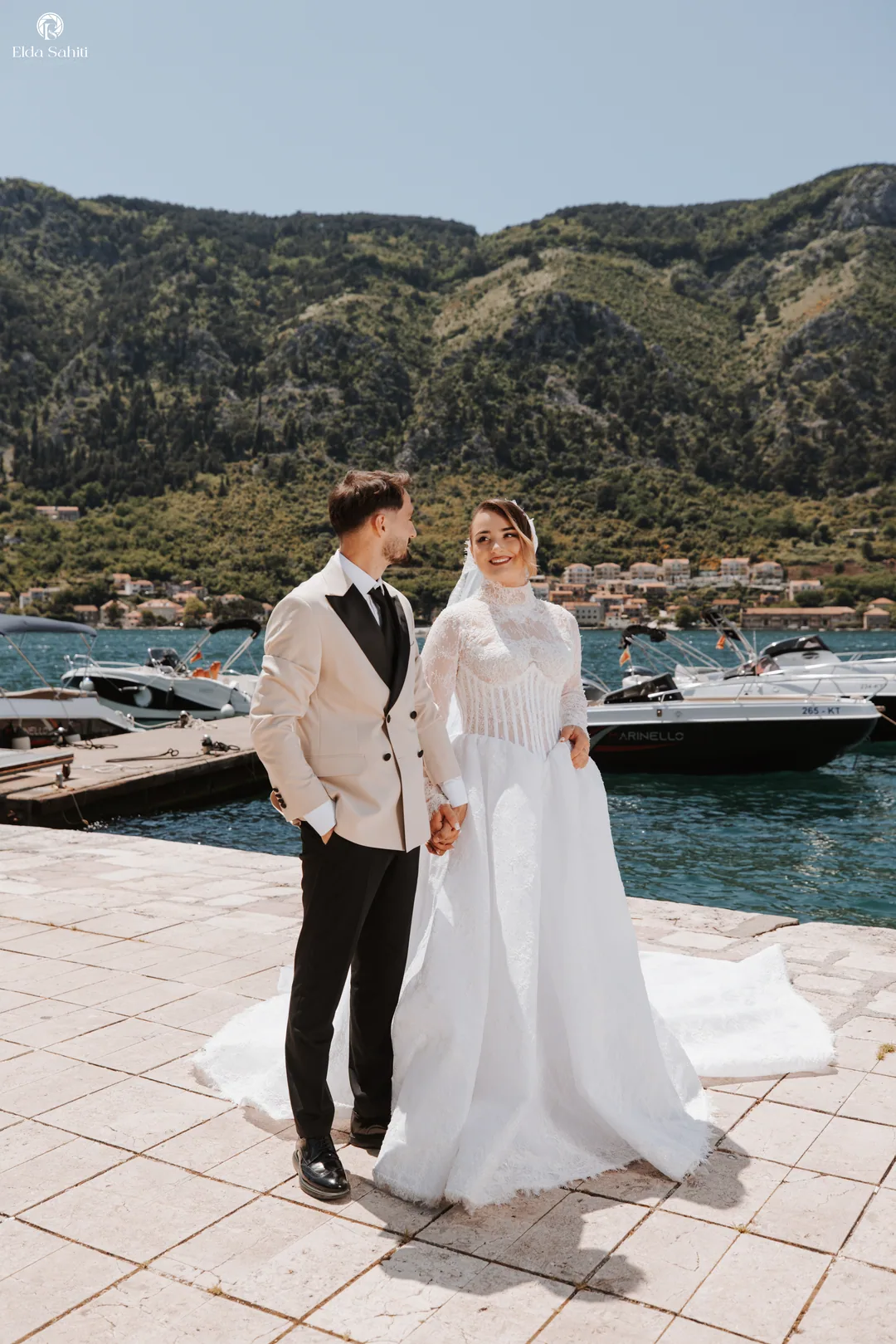 Bride and groom holding hands on a stone quay, boats and mountains behind