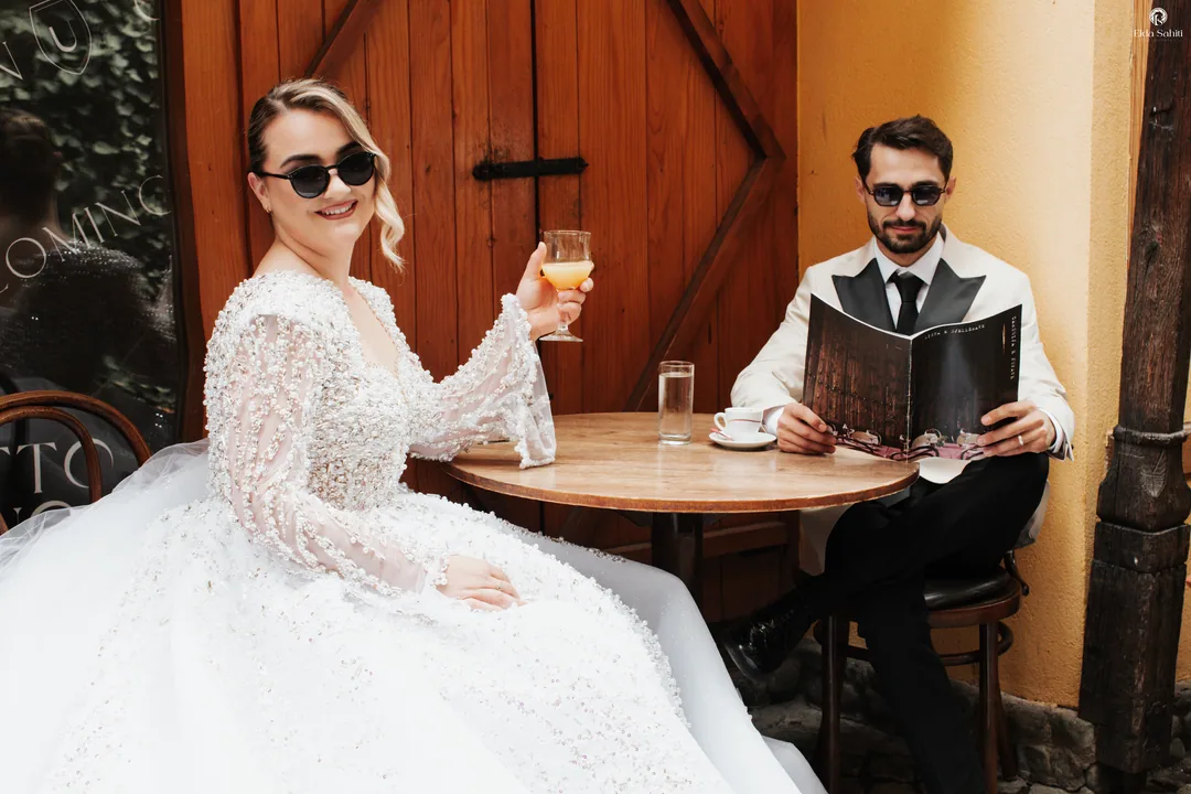 Bride and groom in sunglasses at a café table, her glass raised, a menu in his hands