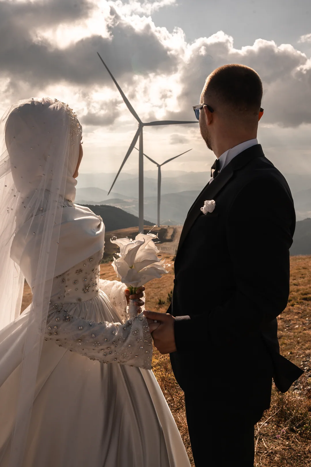 Bride in a hijab and groom looking out at the wind turbines under a cloudy sky