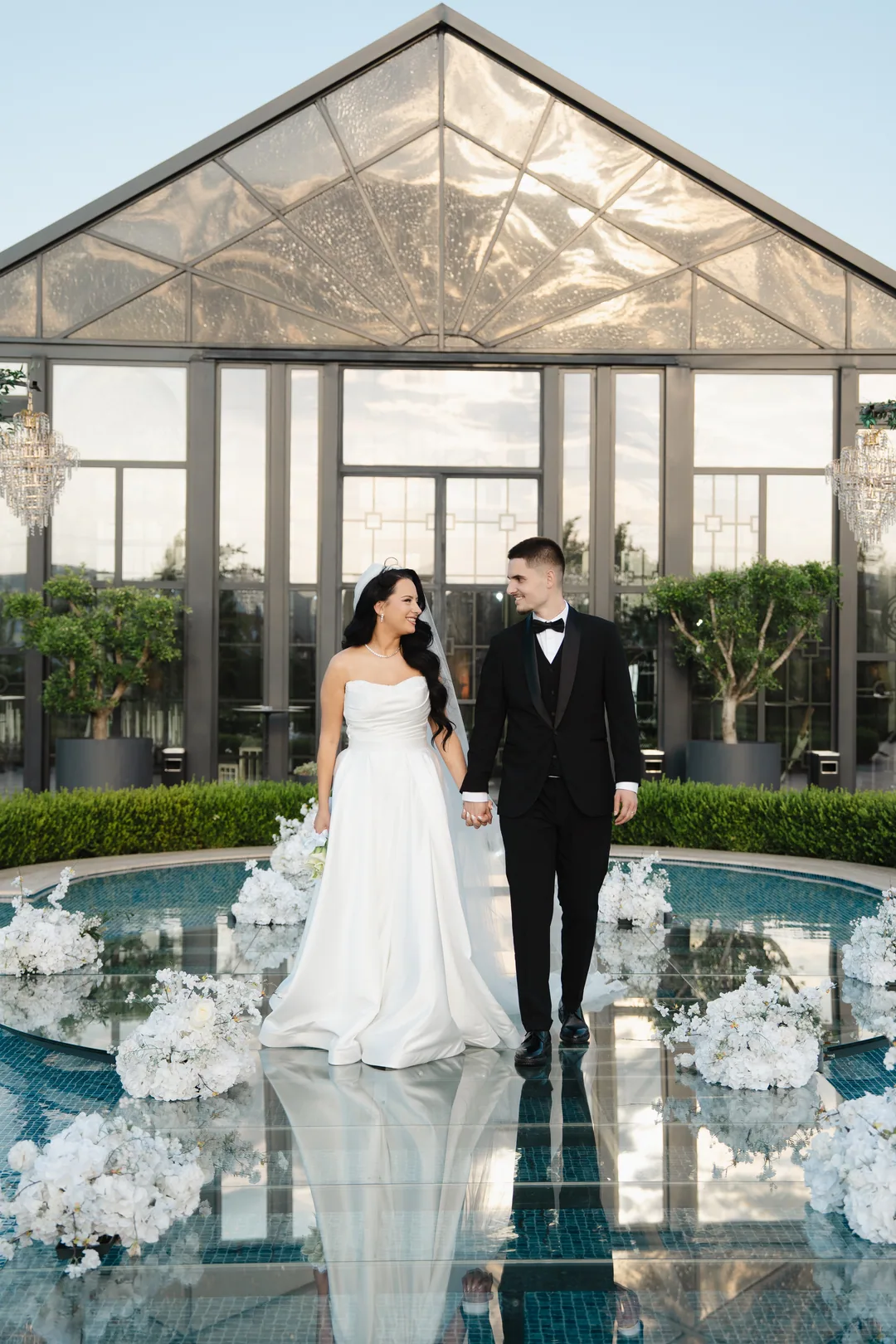 Couple walking hand in hand across a glass walkway over a pool of white flowers
