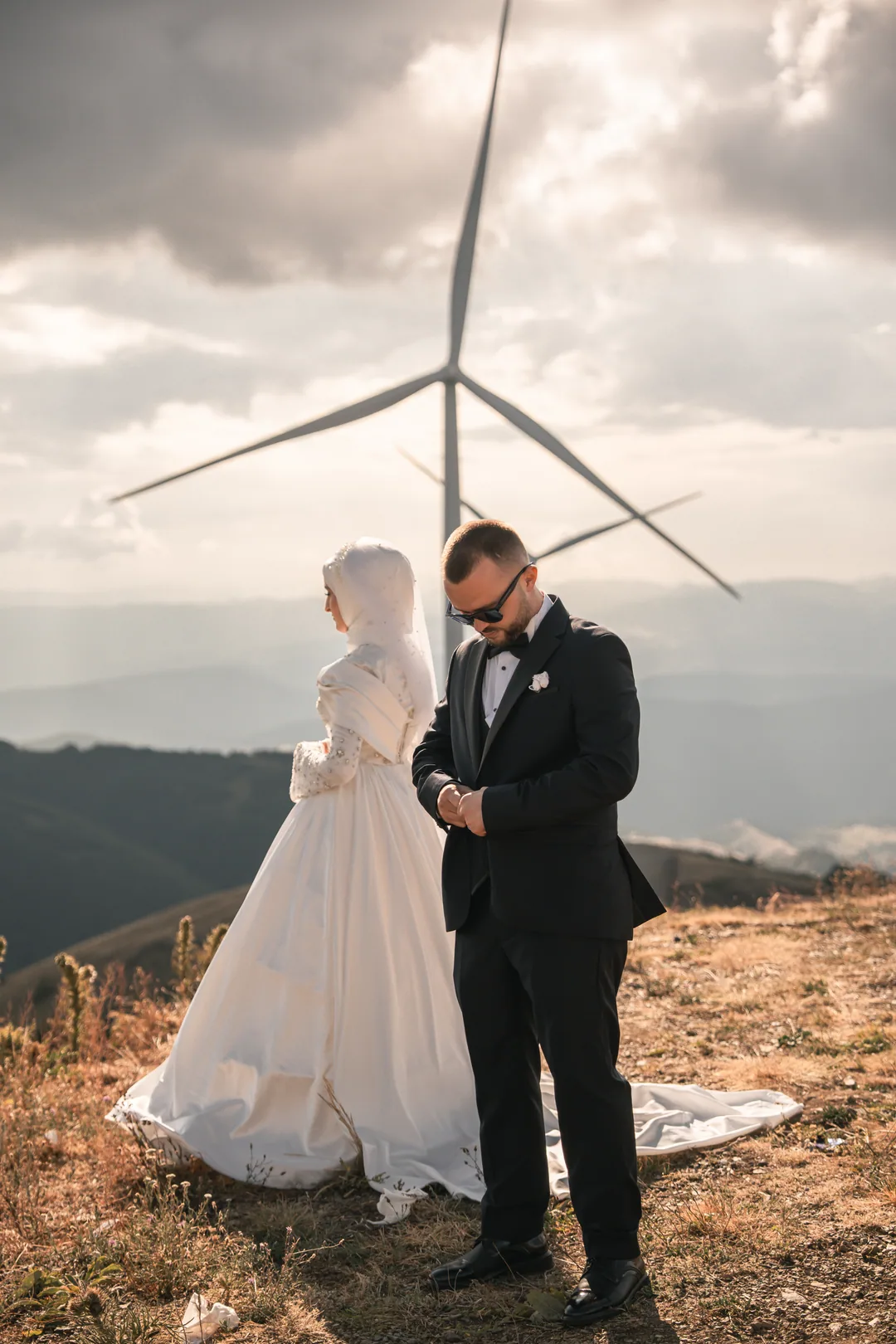 Bride in a hijab and groom on a mountain ridge, a wind turbine behind them