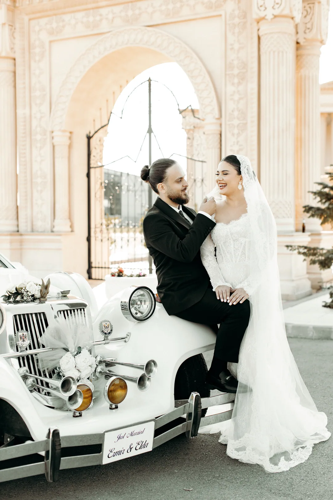 Couple sitting on a white vintage car in front of an ornate carved gate