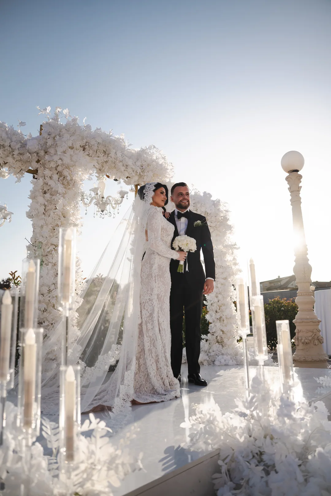 Bride and groom under a white floral arch at sunset, candles on either side