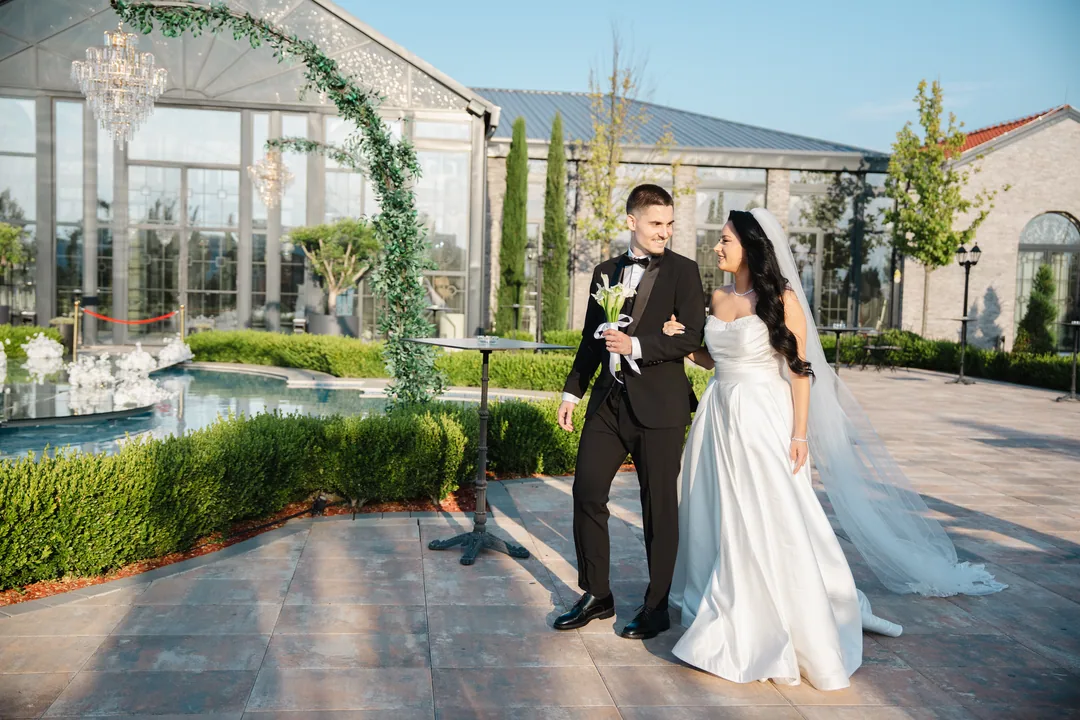 Bride and groom walking under a green arch, a fountain behind them