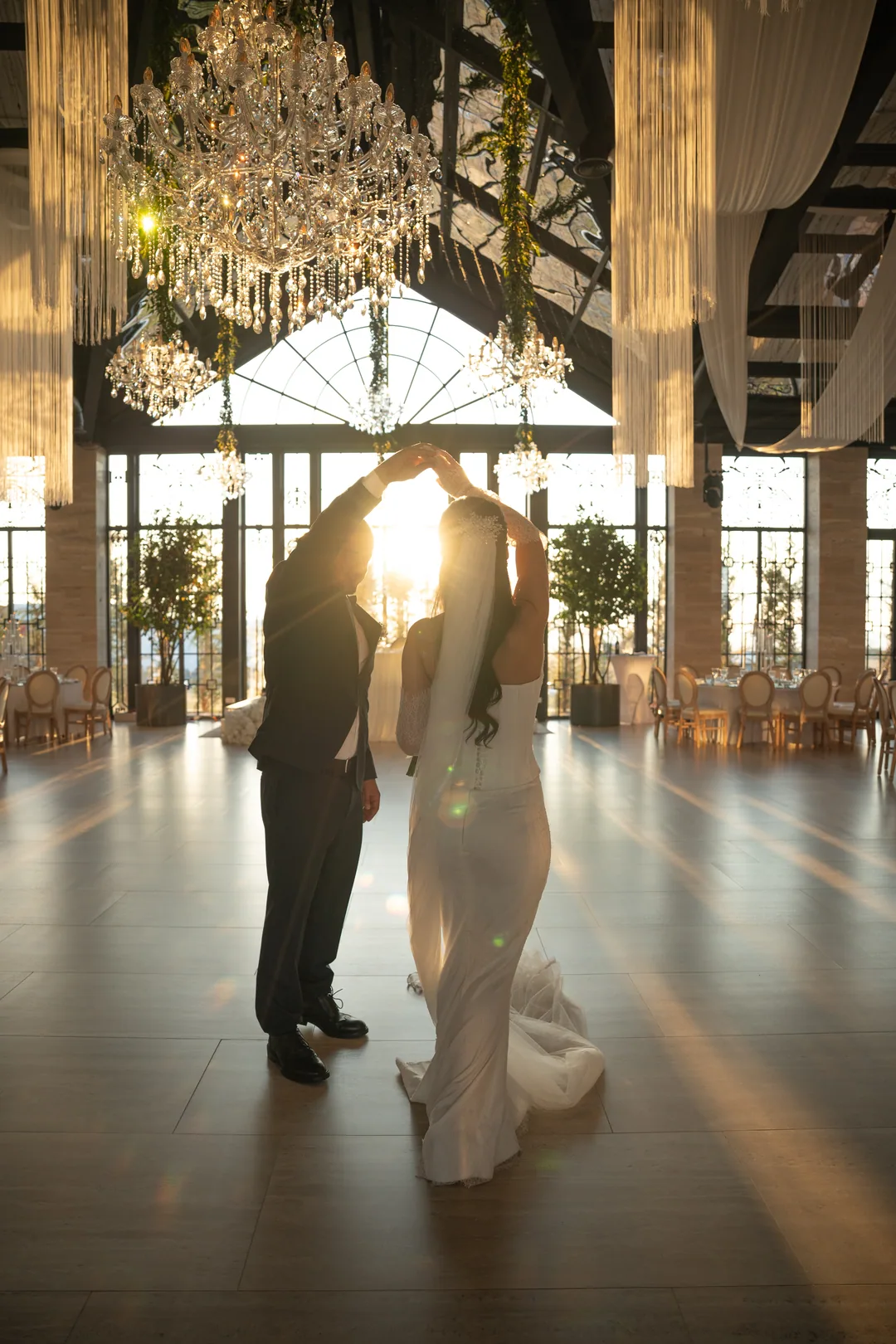 Couple twirling in an empty ballroom, silhouetted against sunlit windows under crystal chandeliers