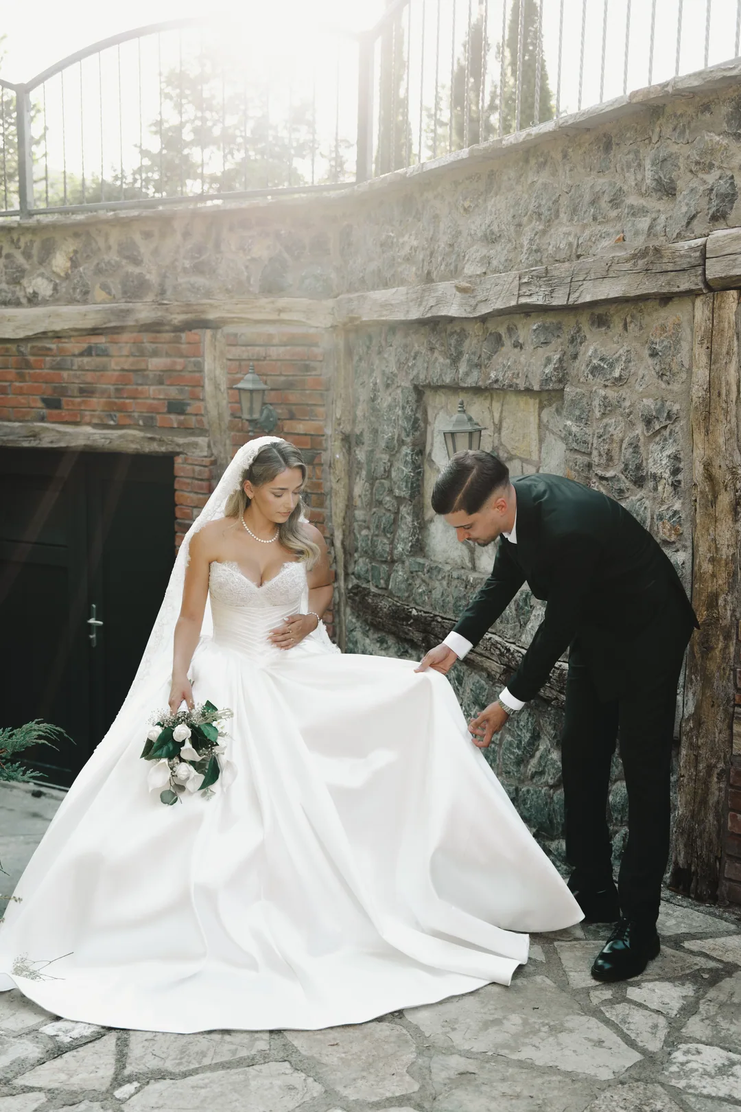 Groom bending to arrange the bride's skirt in front of a stone wall