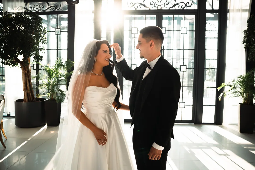 Groom brushing back the bride's veil in a bright hall with tall windows