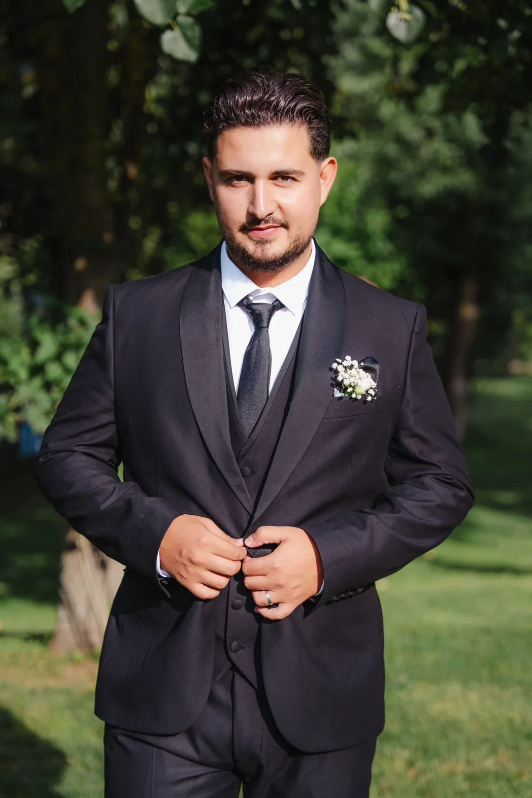 Groom buttoning the jacket of his three-piece suit under the trees