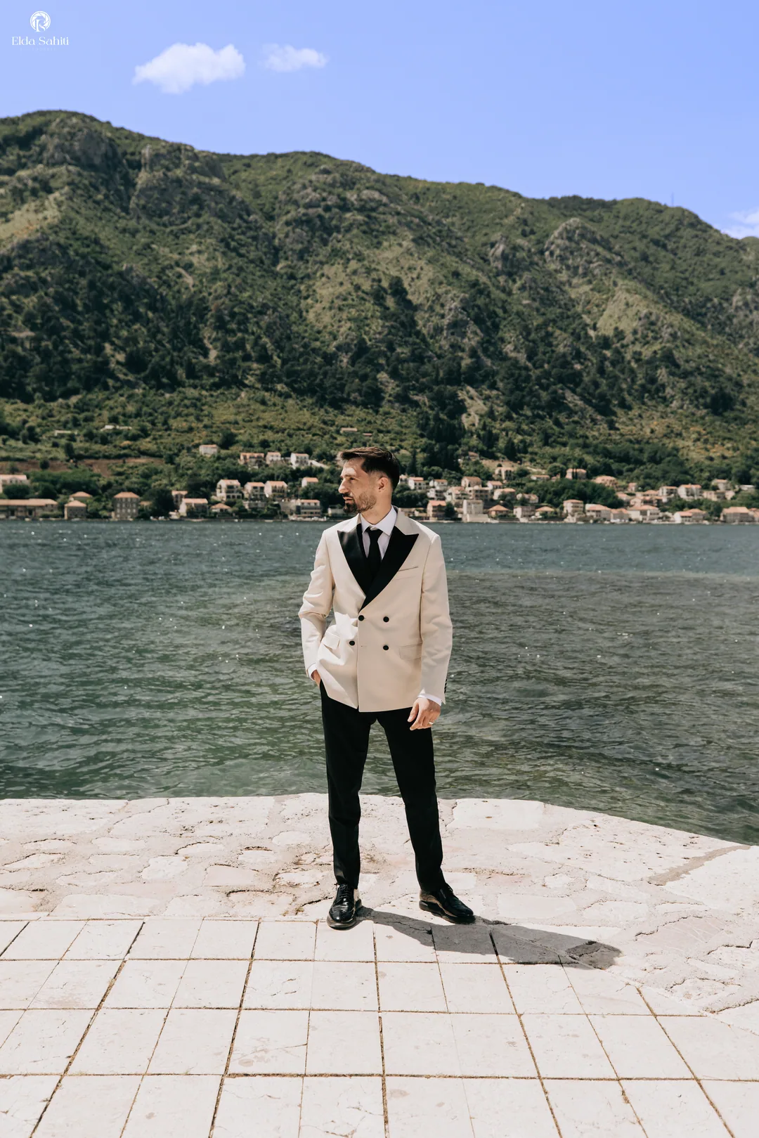 Groom in a cream double-breasted jacket on the quay, the bay and mountains behind