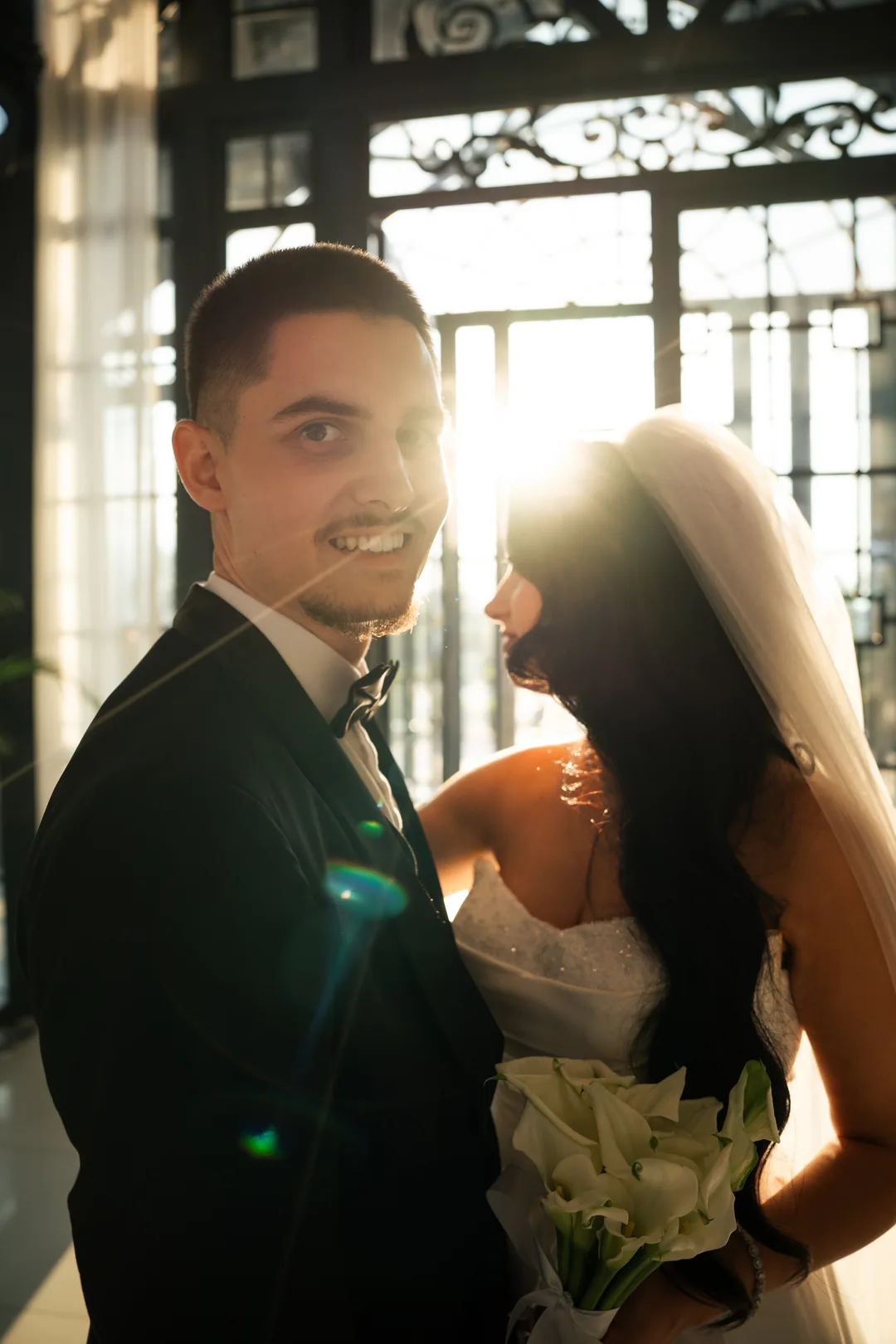 Groom grinning at the camera as the sun flares past the bride's veil