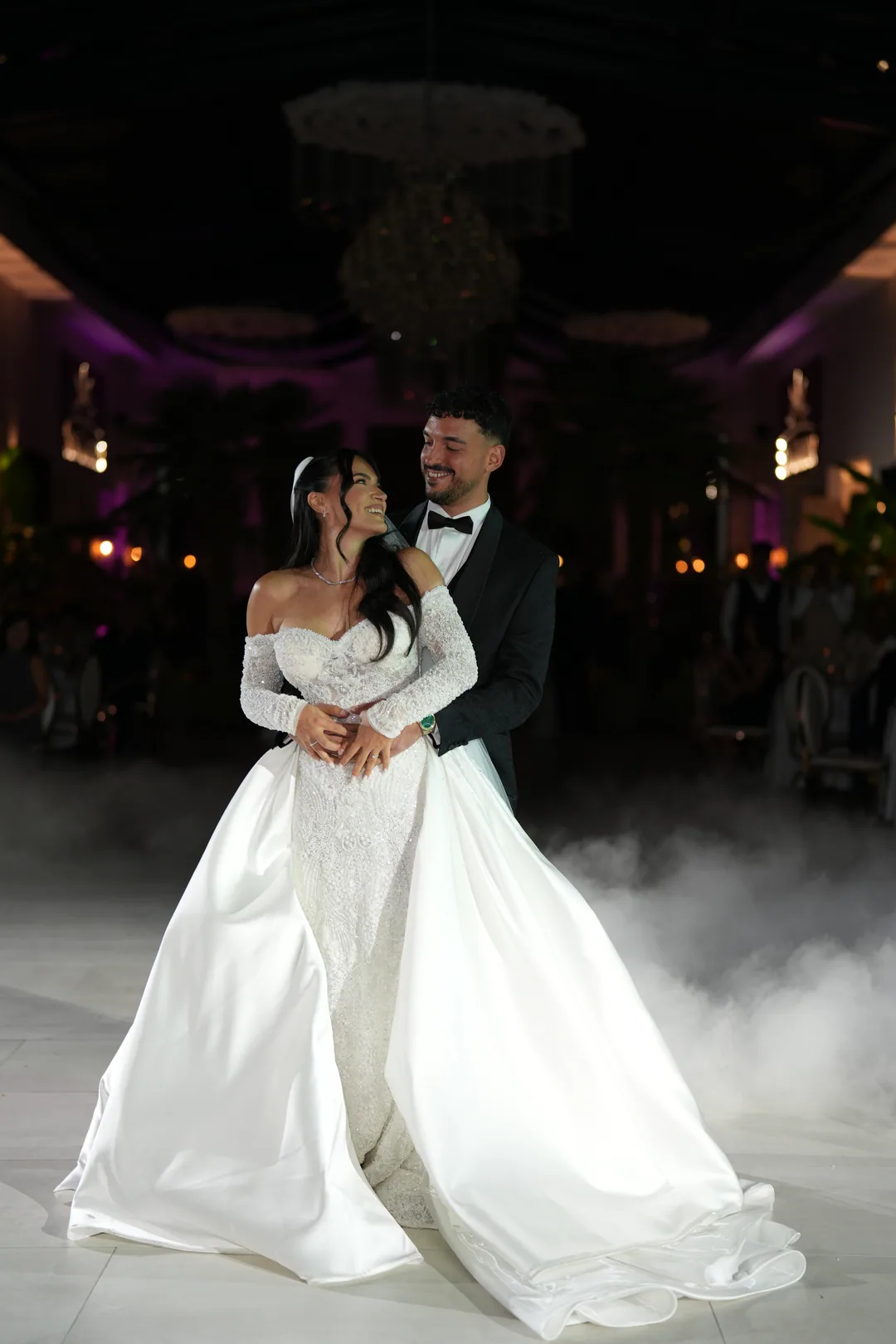 Groom holding the bride from behind on the dance floor, her satin train spread in the fog