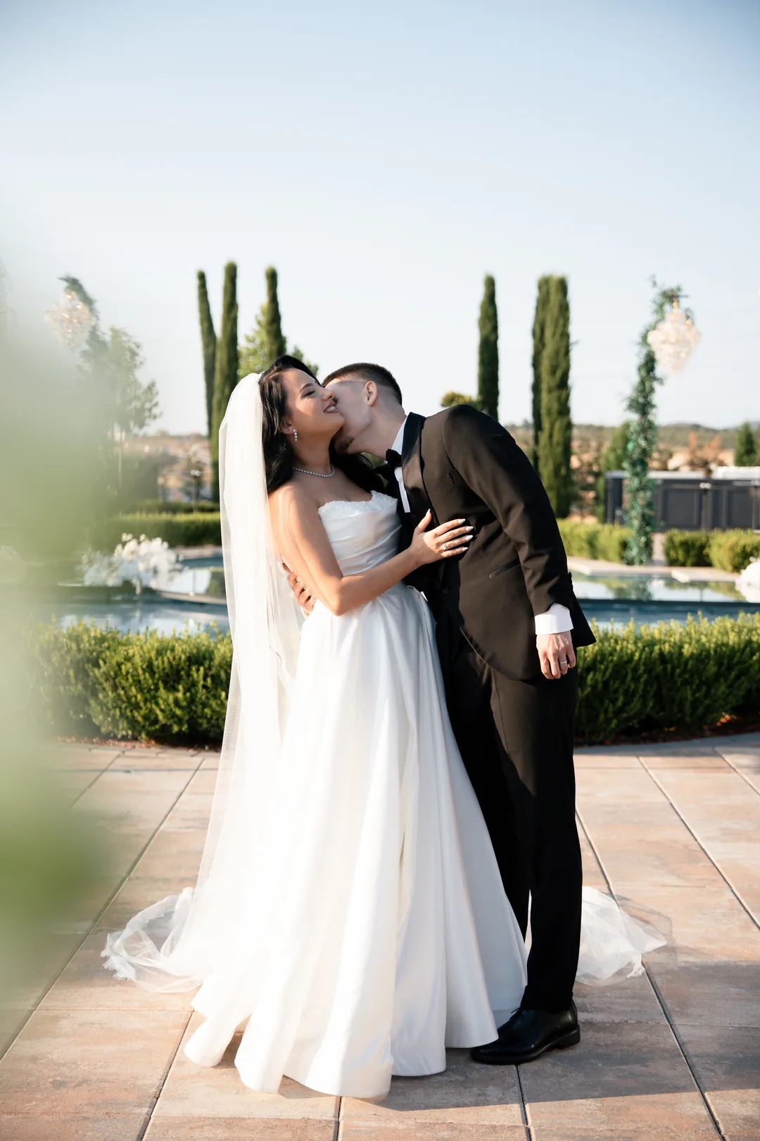 Groom kissing the bride's cheek in a fountain garden, cypresses behind