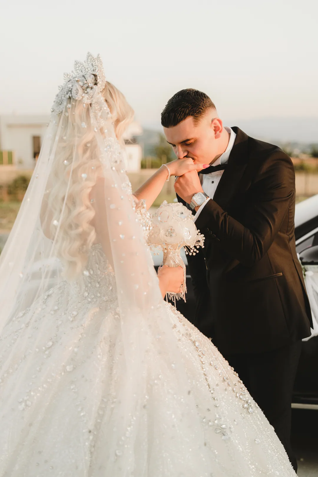 Groom kissing the bride's hand beside the car at golden hour