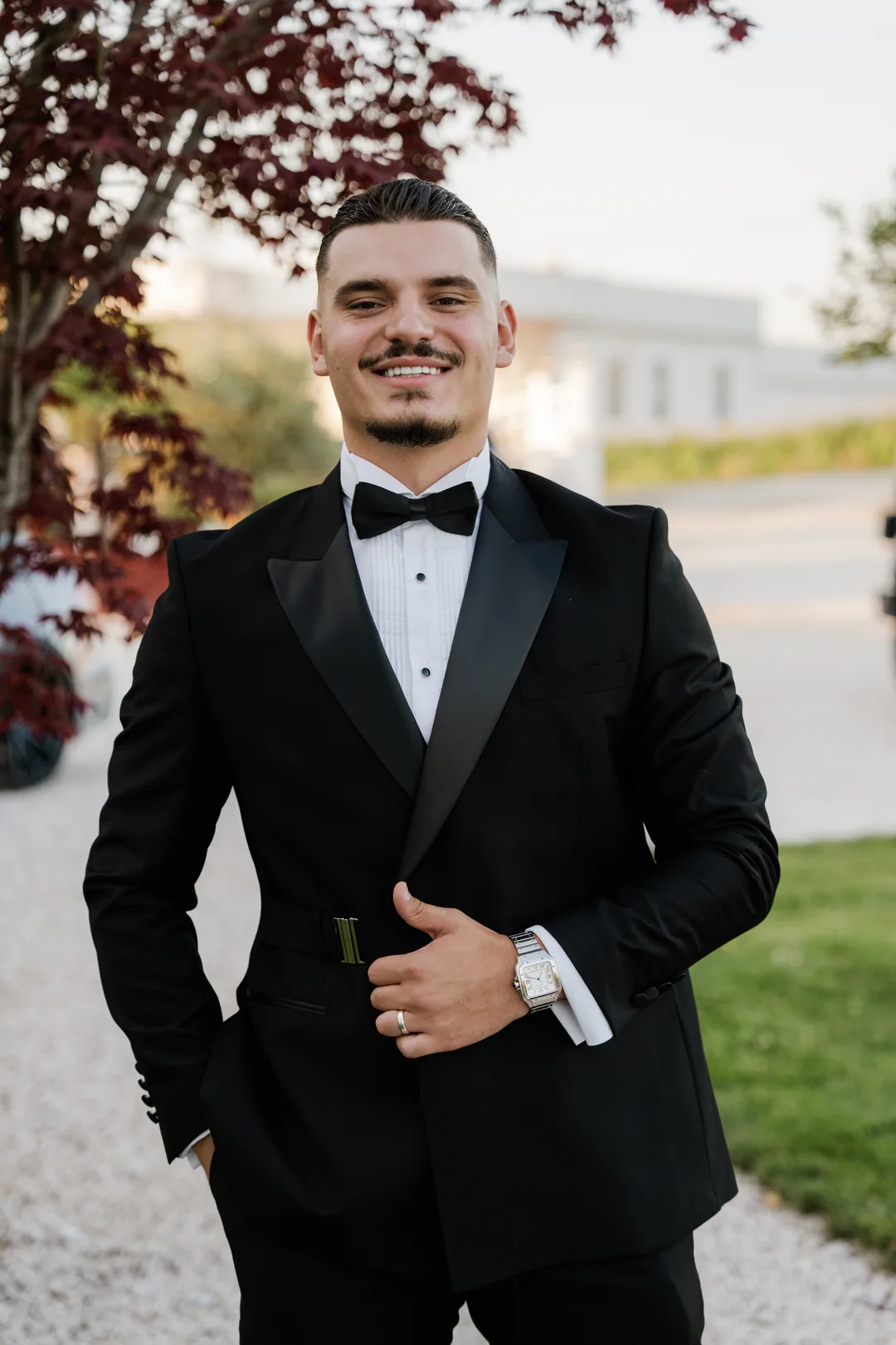 Groom smiling in a black tuxedo, a red-leaved tree behind him