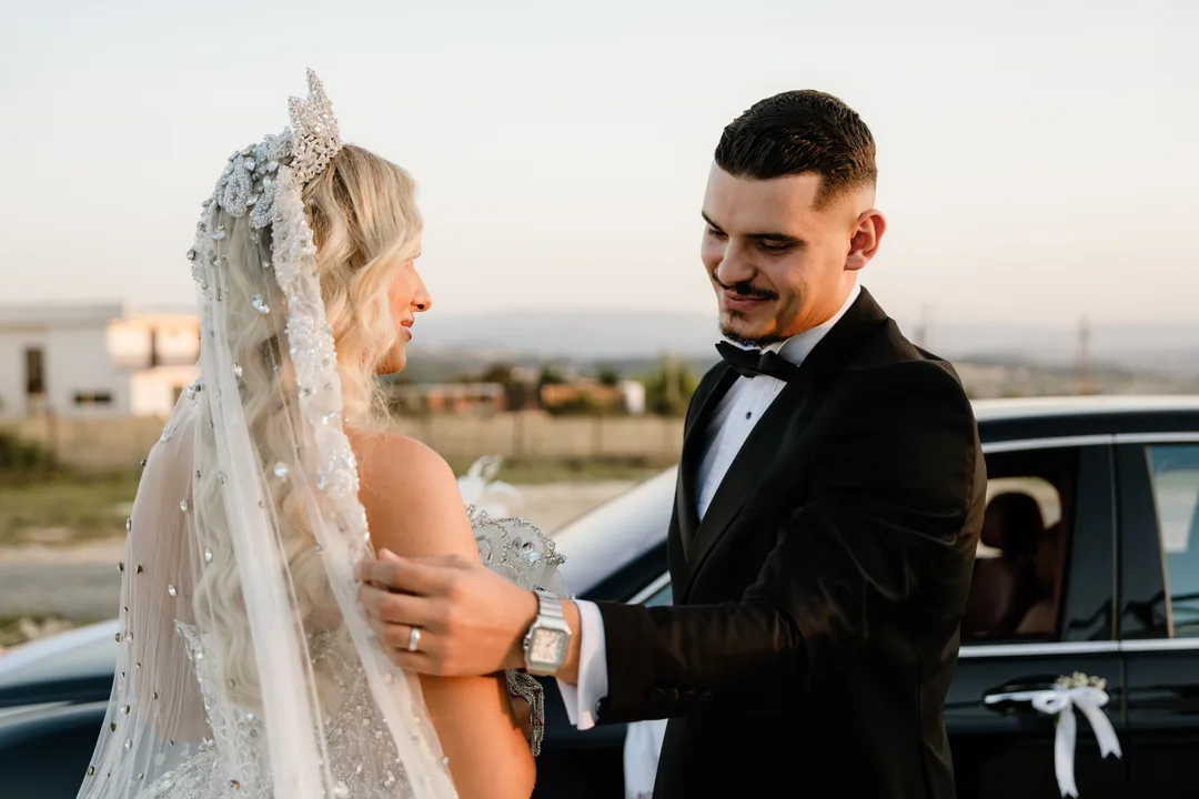 Groom straightening the bride's veil beside the car, both smiling