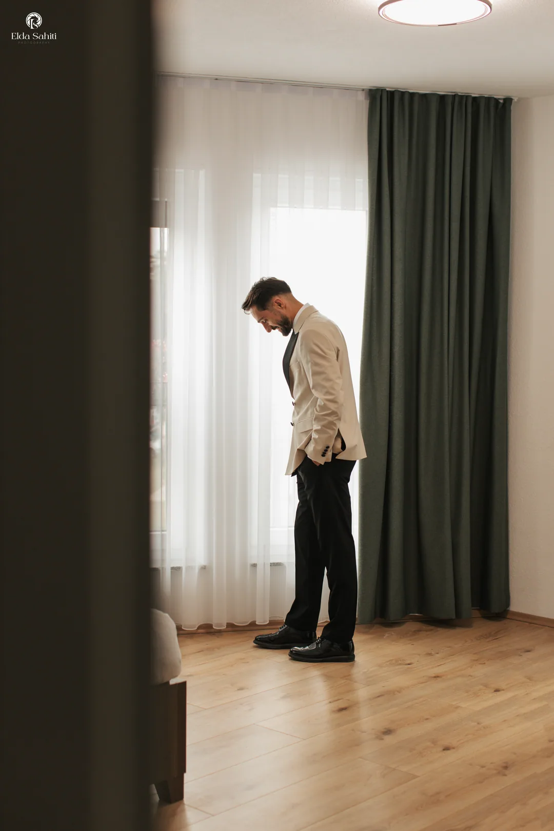 Groom waiting alone by the window in a cream jacket, hands in his pockets