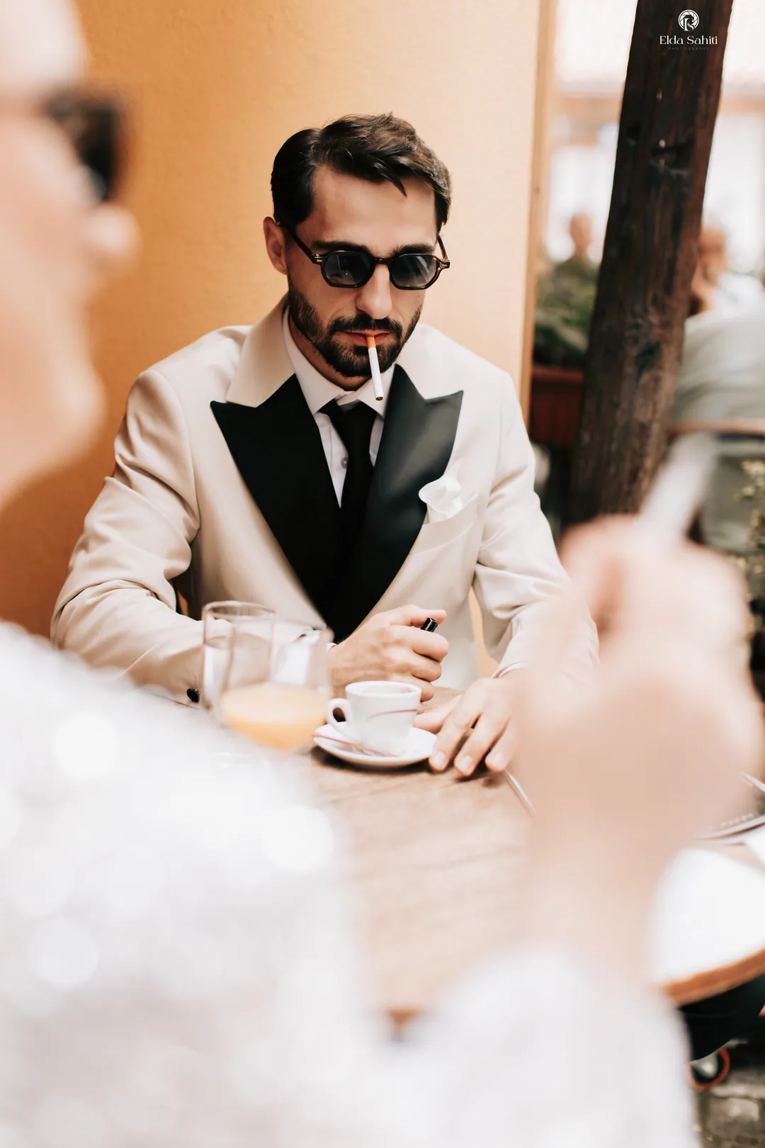 Groom in a cream tuxedo and sunglasses, a cigarette between his lips, at the café table
